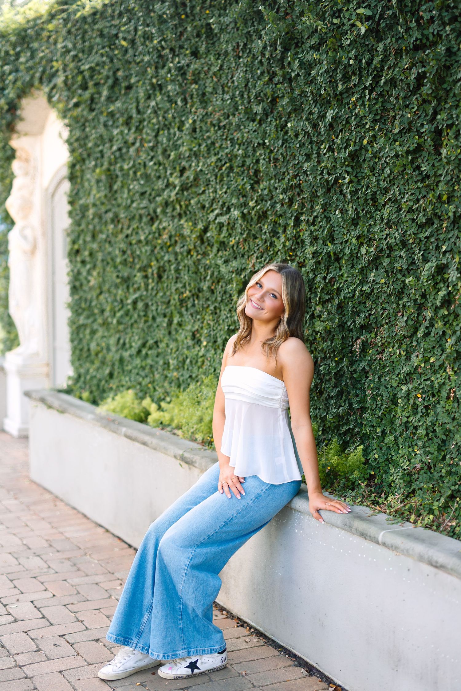 Young woman in white tube top and wide-leg jeans sitting on a stone ledge against a lush green ivy-covered wall.