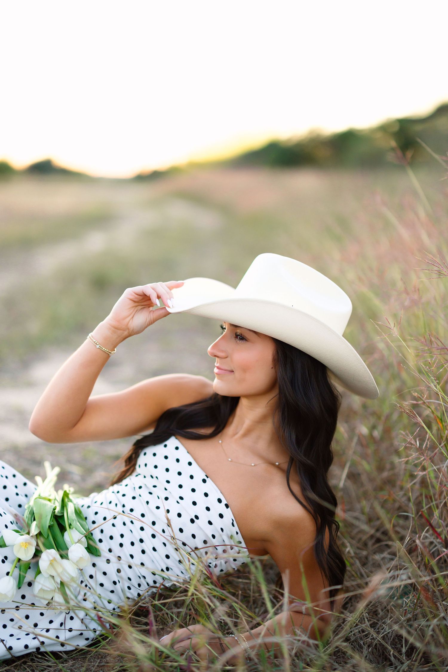 A person in a polka dot dress and white hat poses gracefully in a field of wildflowers during golden hour.