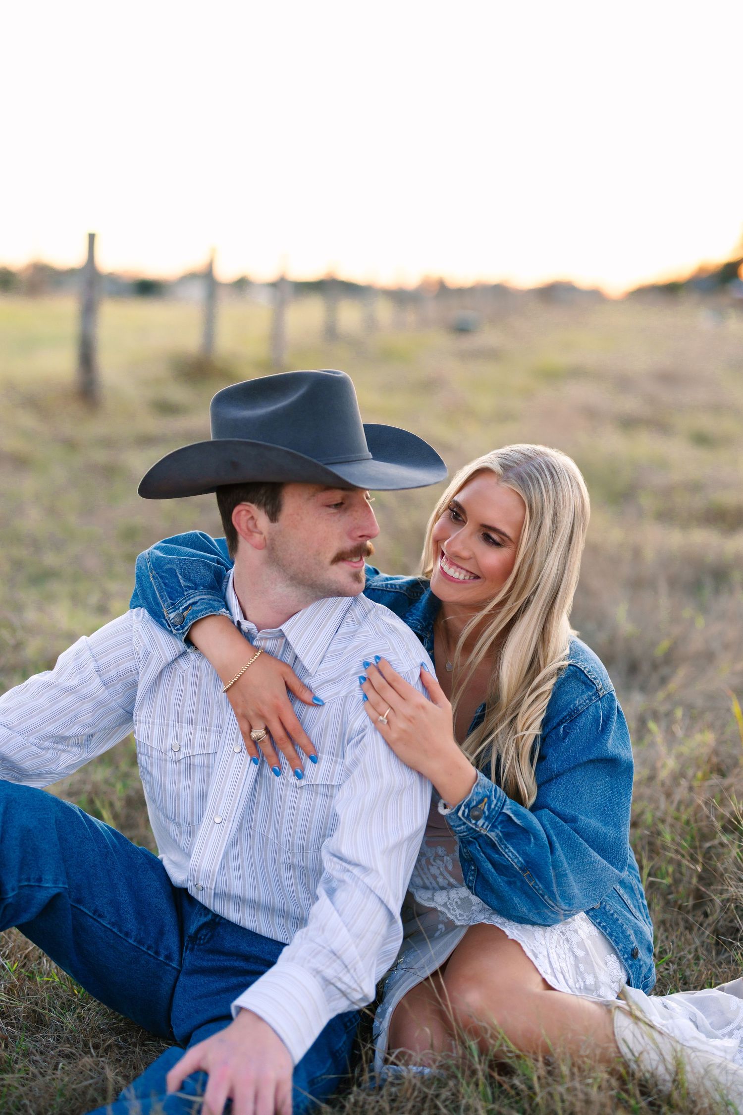 Couple sitting together in a golden field, woman in denim jacket smiling and embracing man in cowboy hat.