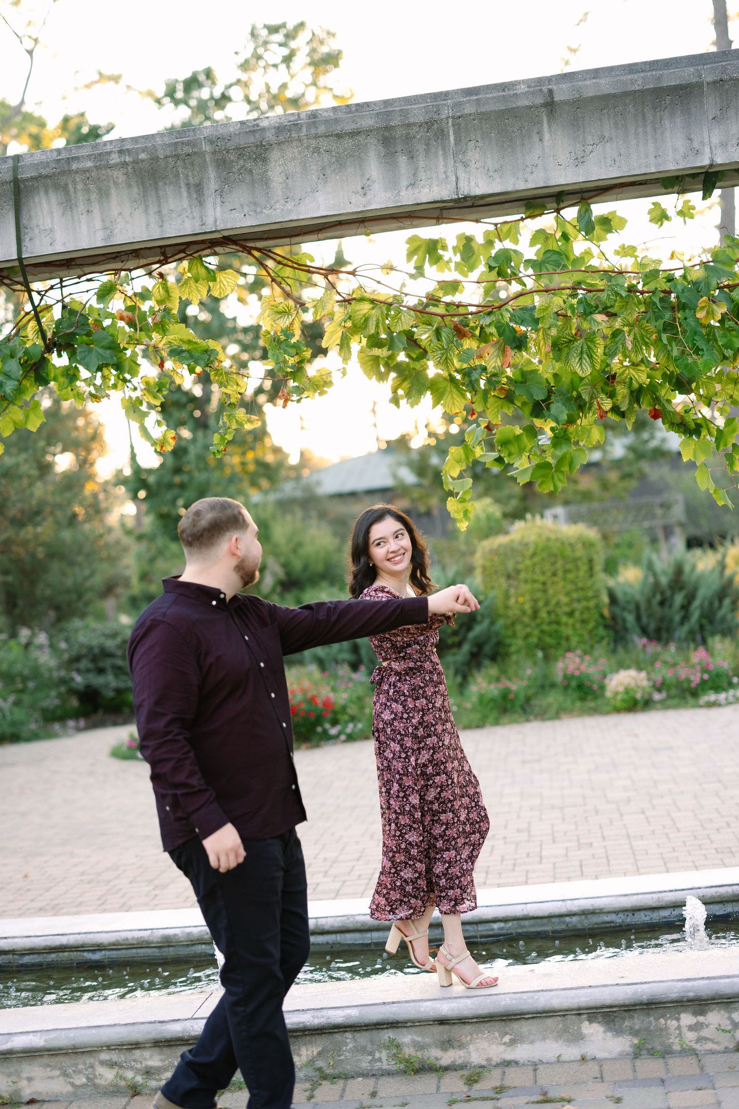 Couple dancing outdoors near a fountain, surrounded by lush greenery, woman in floral dress smiling at partner.