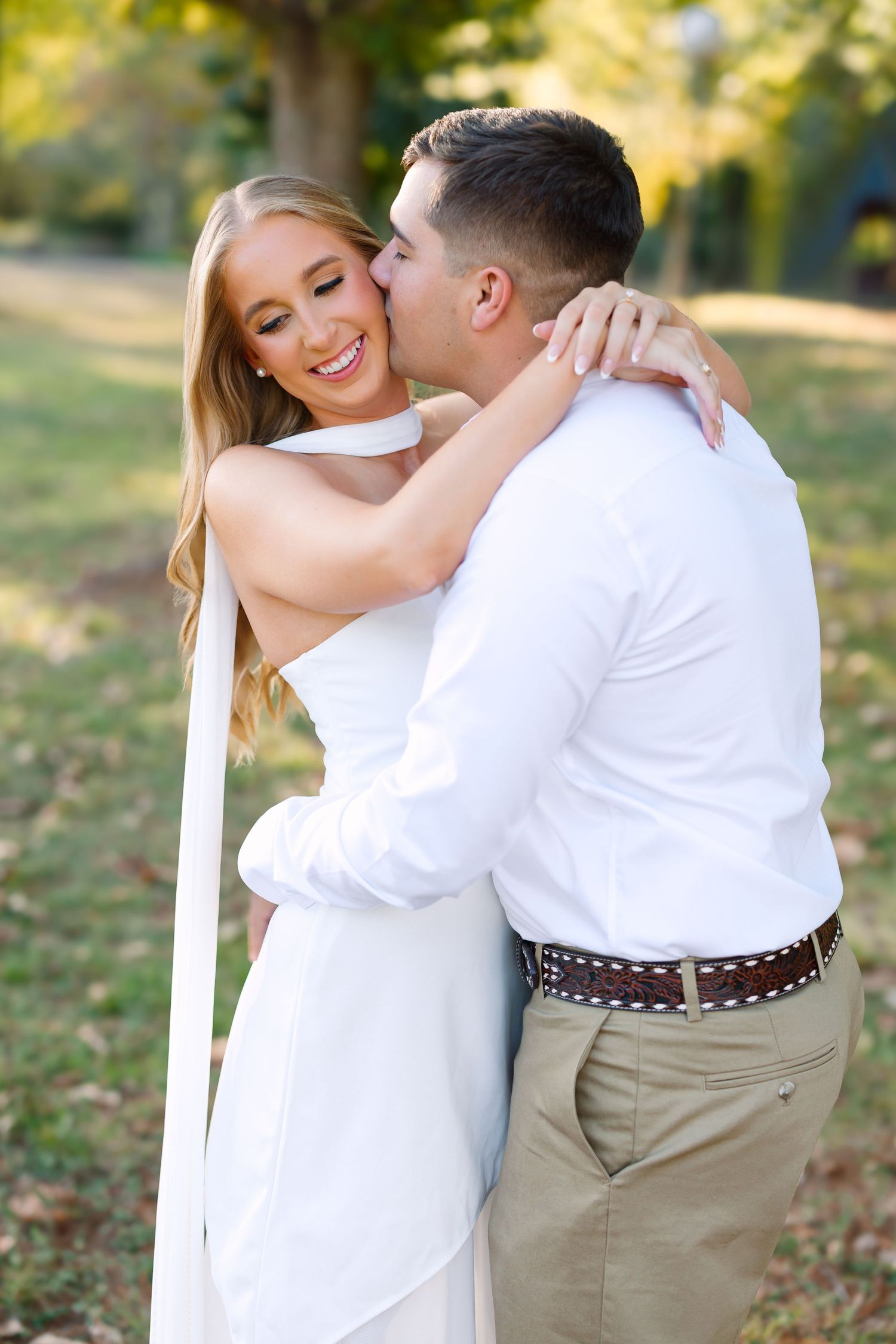 Groom kissing smiling bride on cheek outdoors, both wearing white with green trees in sunny background.