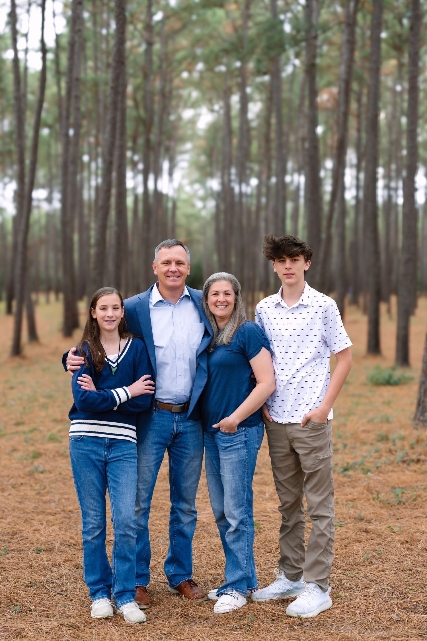 Family of four smiling together in a pine forest, parents with teenage son and daughter in casual attire.