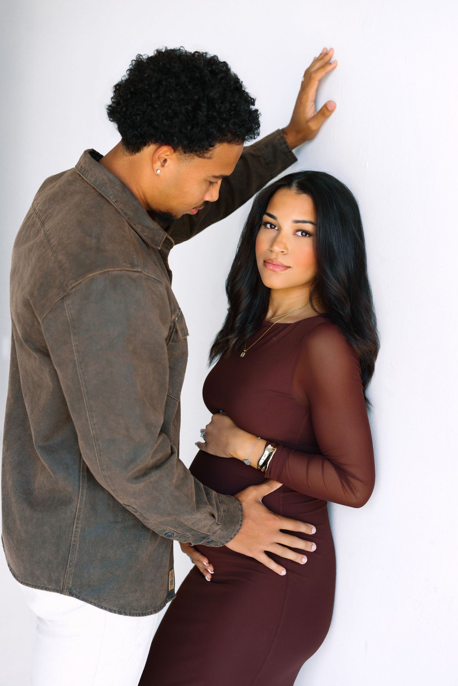 A couple poses tenderly against a white wall, the woman in a burgundy dress cradling her baby bump.