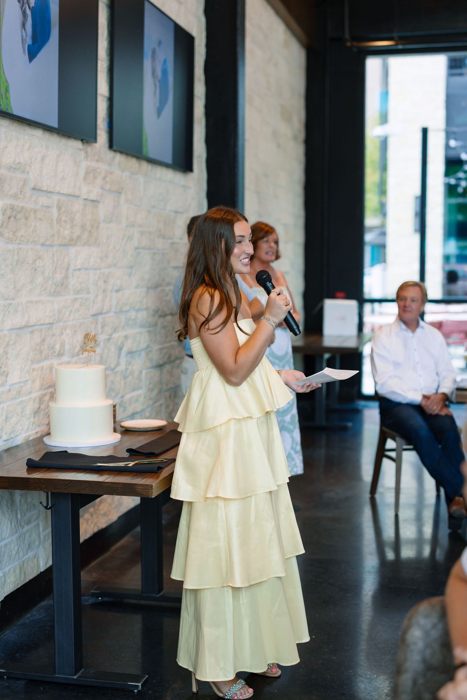 Young woman in yellow tiered dress giving speech at wedding reception with white cake nearby.