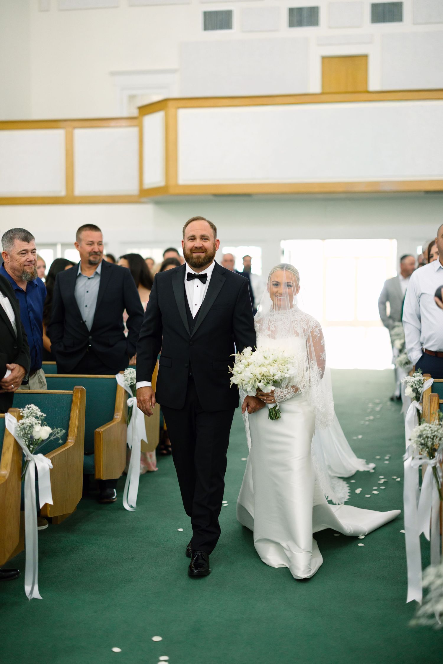Bride in white lace veil and gown walks down church aisle with escort in black tuxedo during wedding ceremony.