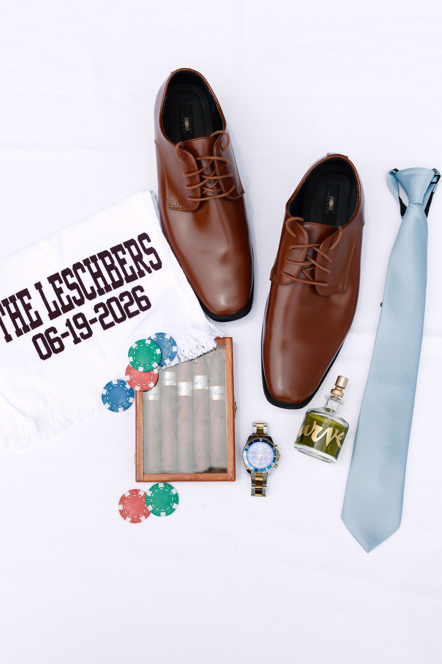 Groom wedding flat lay with brown dress shoes, blue tie, cologne, watch, cigar case, poker chips, and personalized shirt.