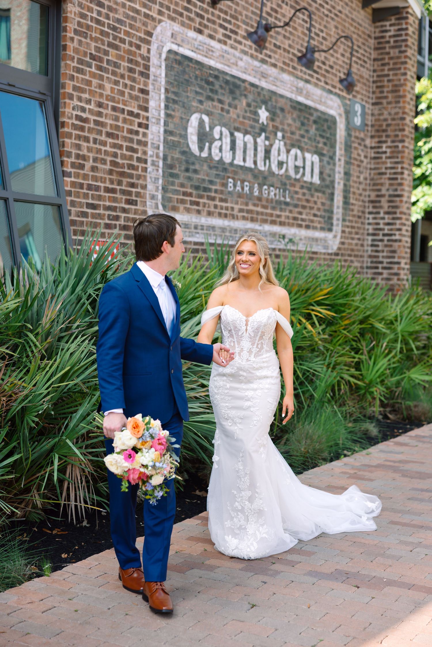 Bride and groom hold hands outside Canteen Bar and Grill, she in a fitted lace gown, he in a blue suit.