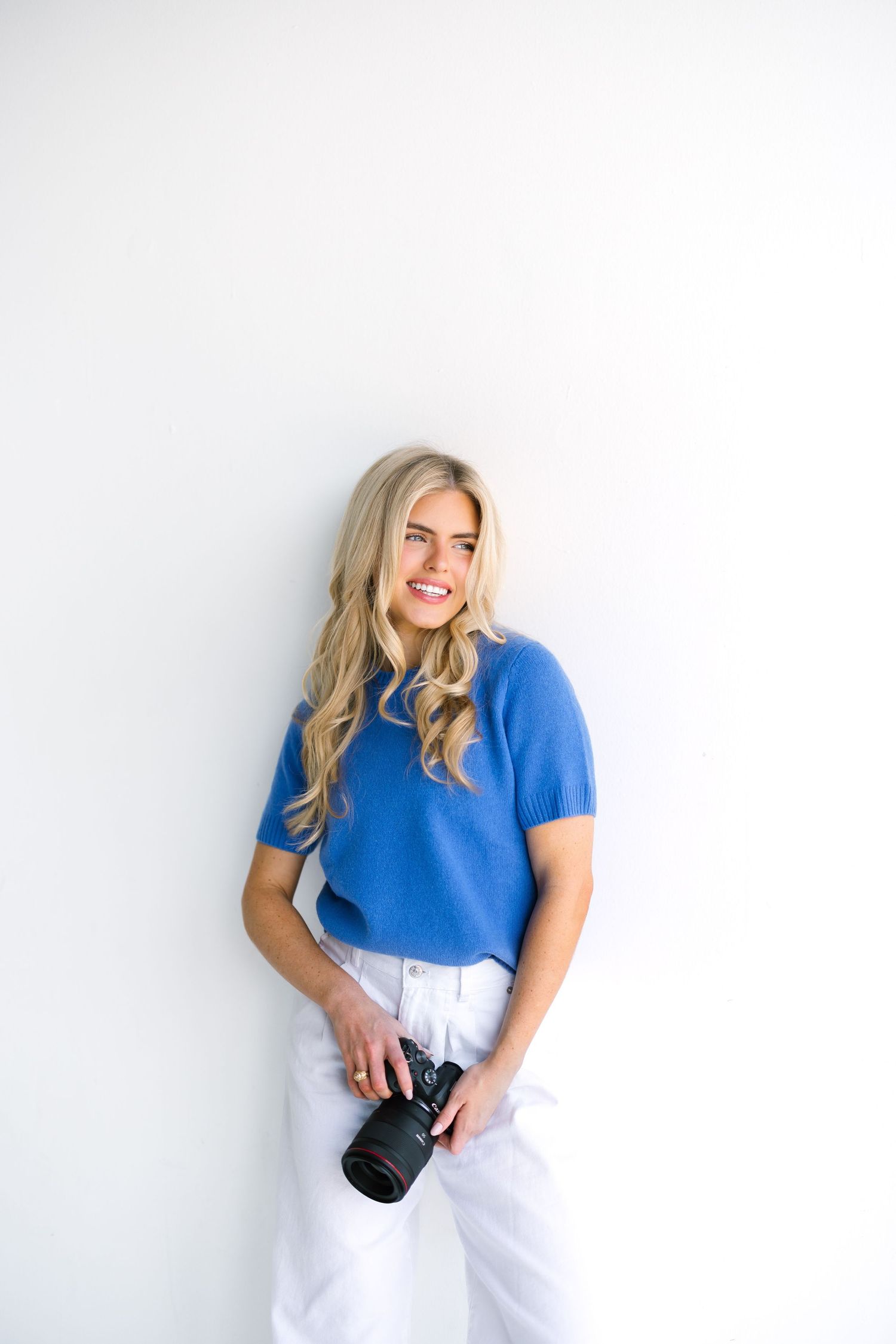 Professional photographer wearing bright blue shirt and white pants poses with camera against white backdrop.