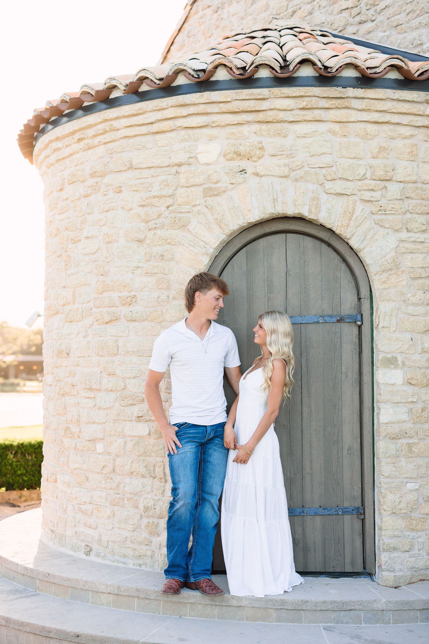 Couple poses together outside stone building with arched wooden door.