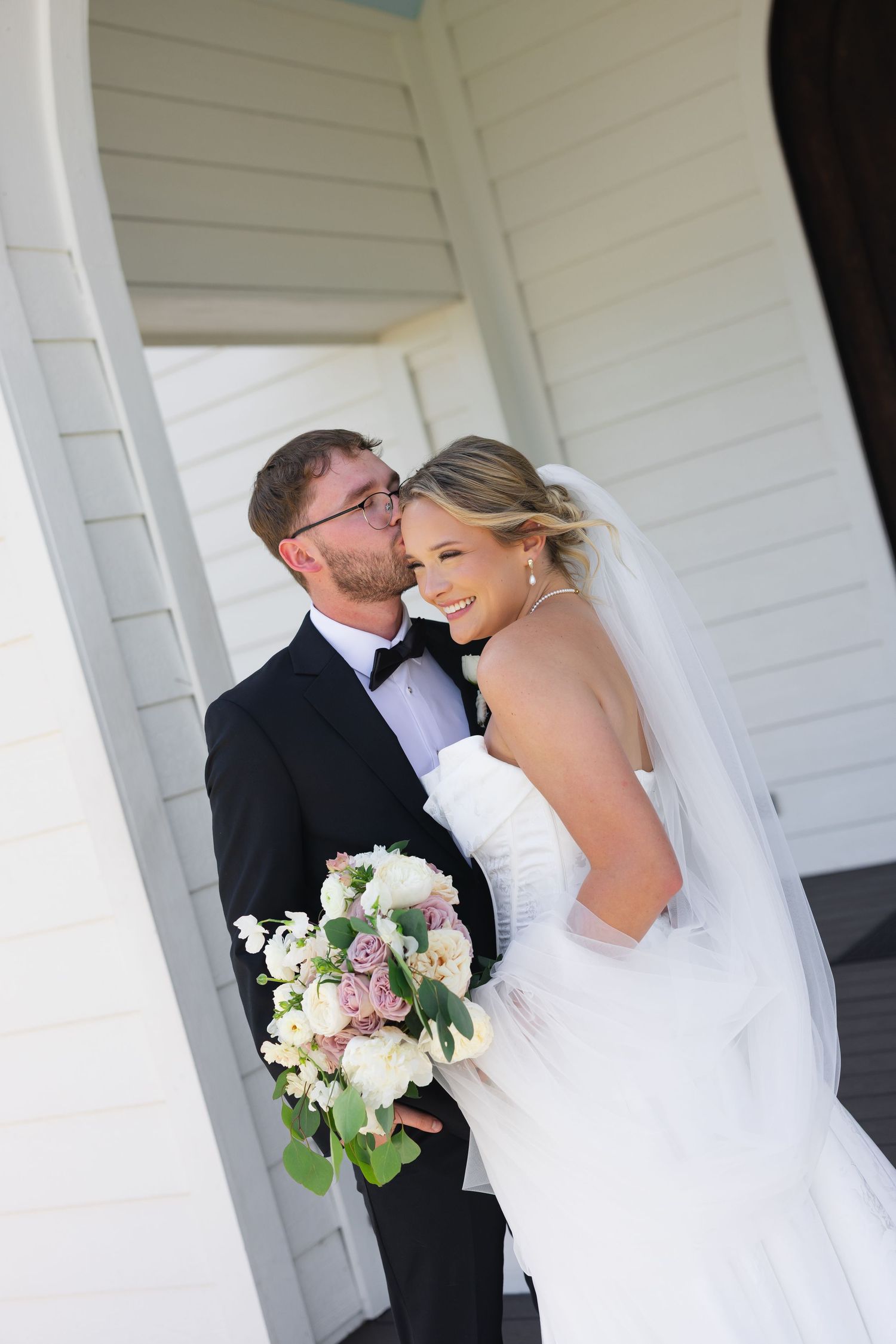Romantic wedding couple moment captured against white building exterior.