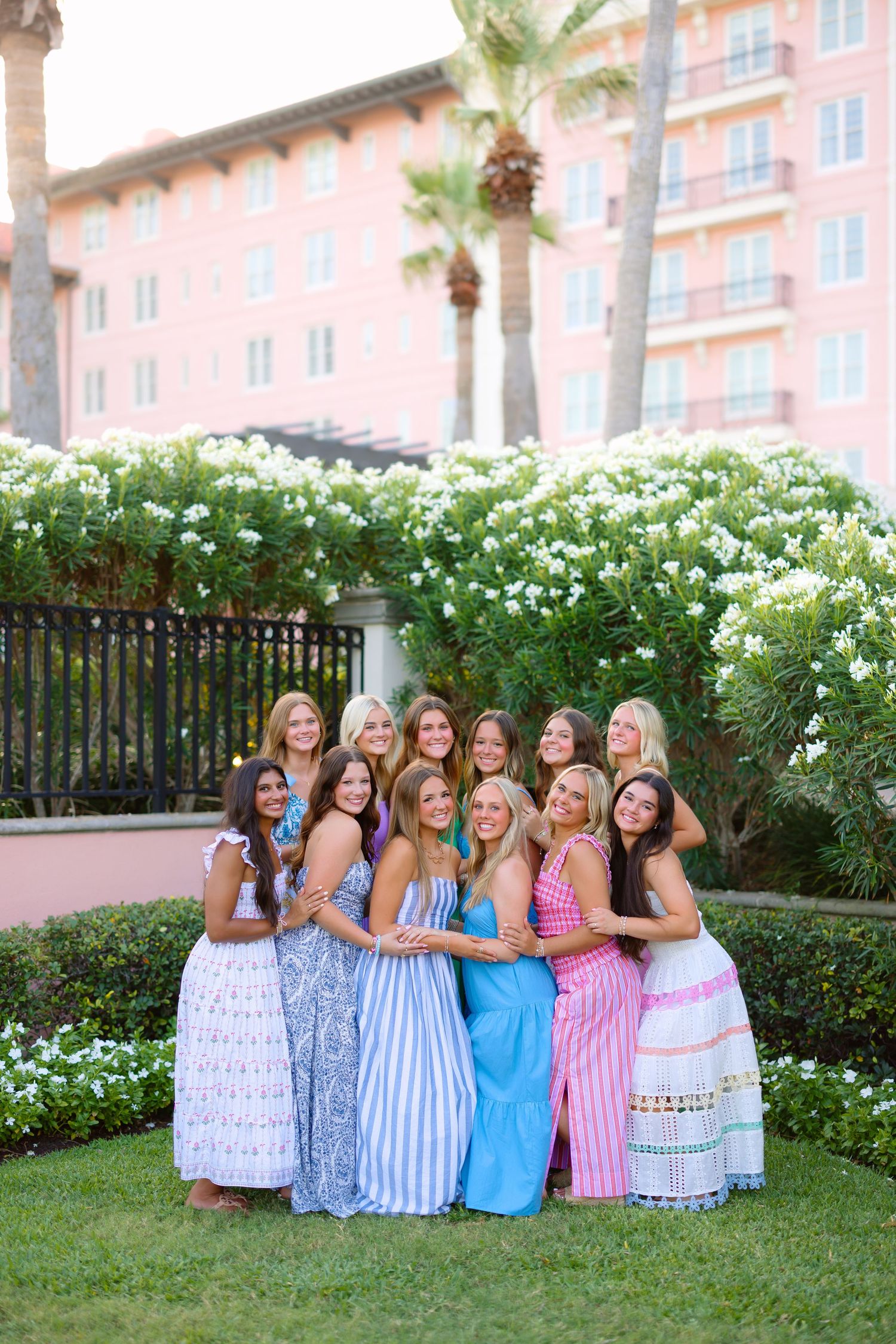 Group of women in colorful maxi dresses posing together outdoors near a pink building with lush white flowers.