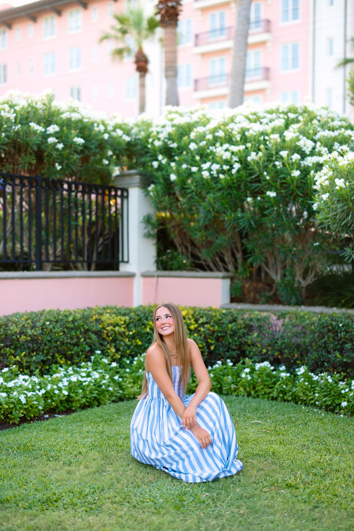 Young woman in blue striped maxi dress sitting on green lawn with white flowering bushes and pink building backdrop.