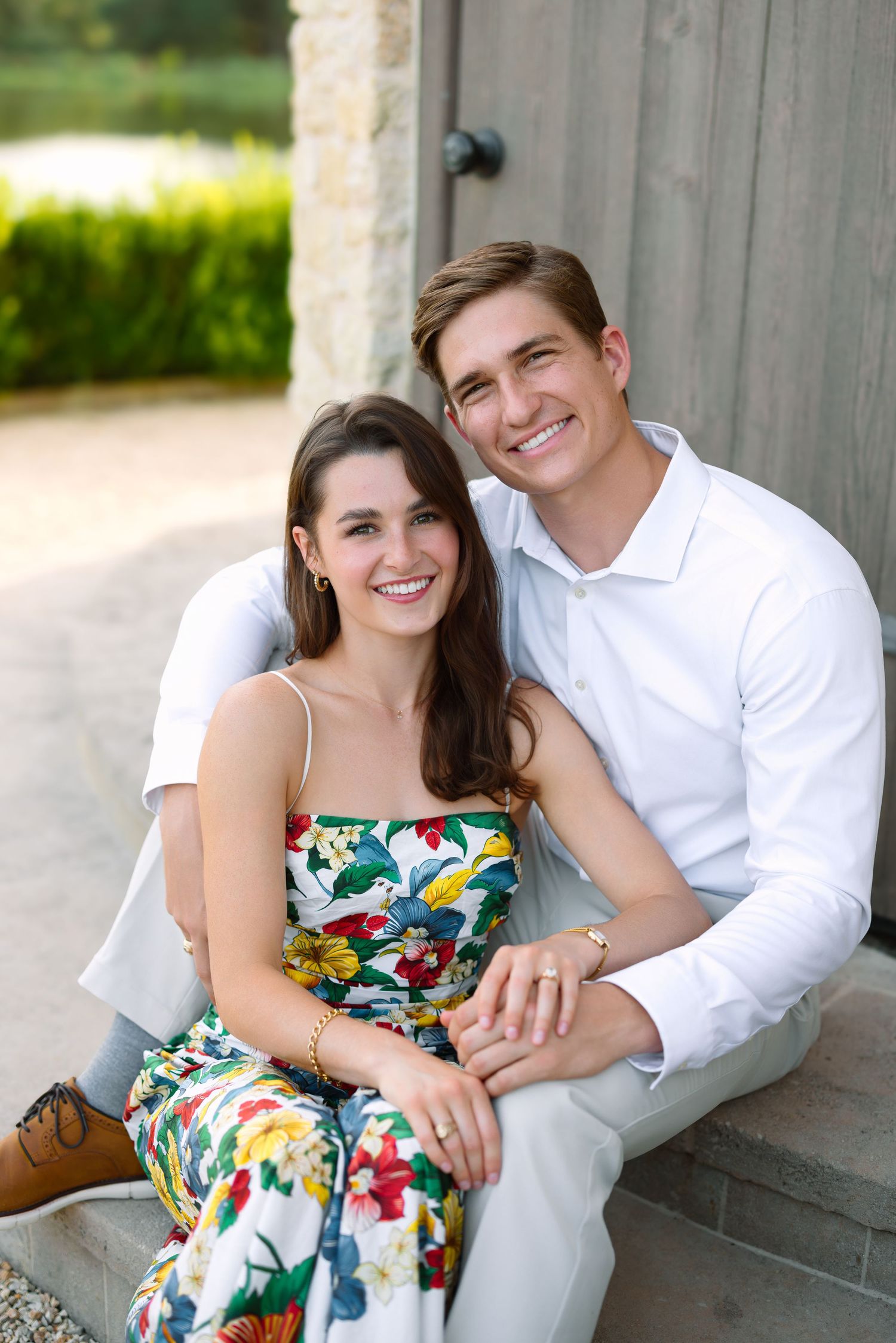 A smiling young couple sits together on steps, the woman in a floral dress and the man in a white shirt, holding hands.