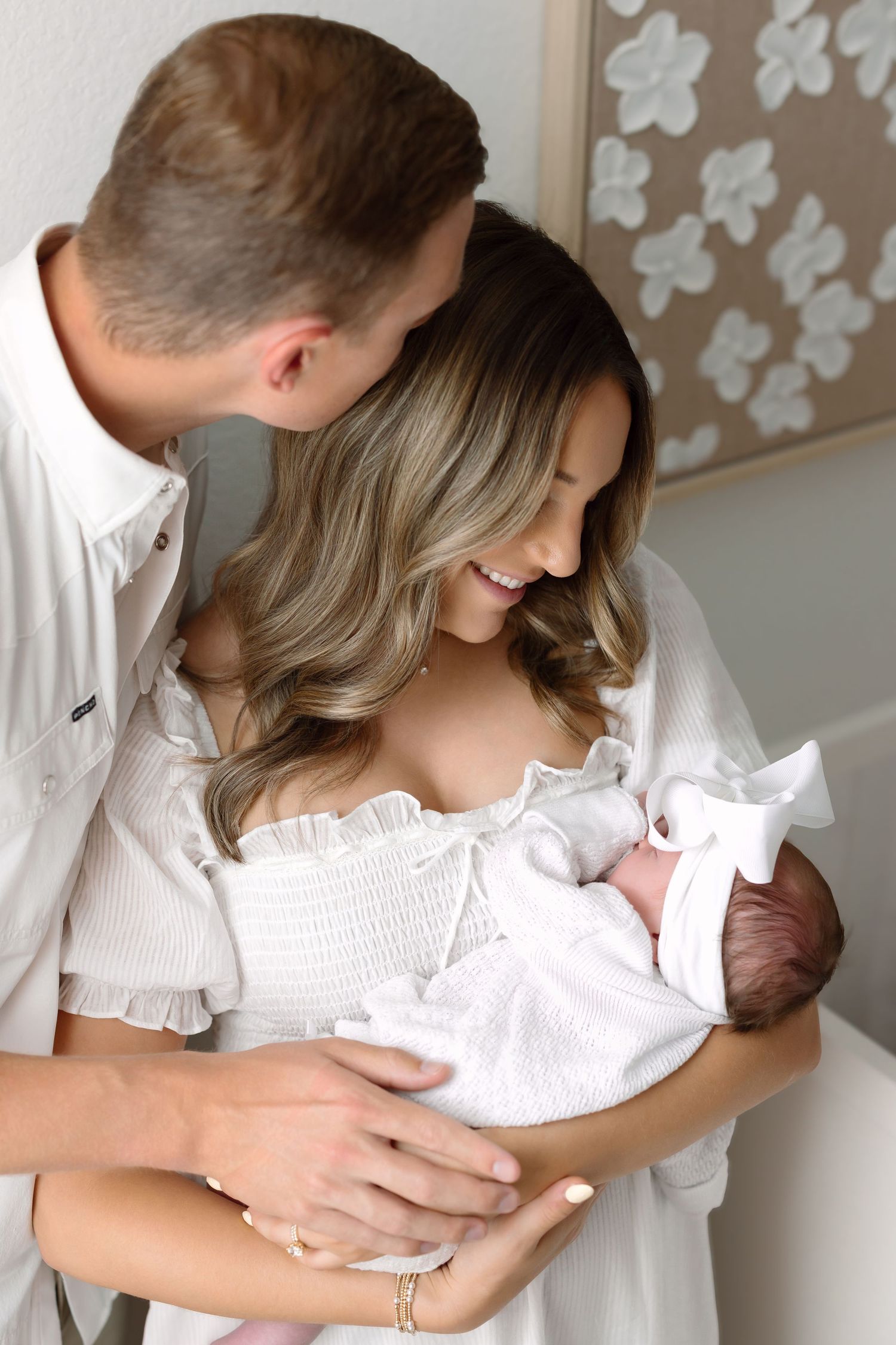 Young couple in white outfits smiling at their newborn baby in a bright, elegant nursery setting.