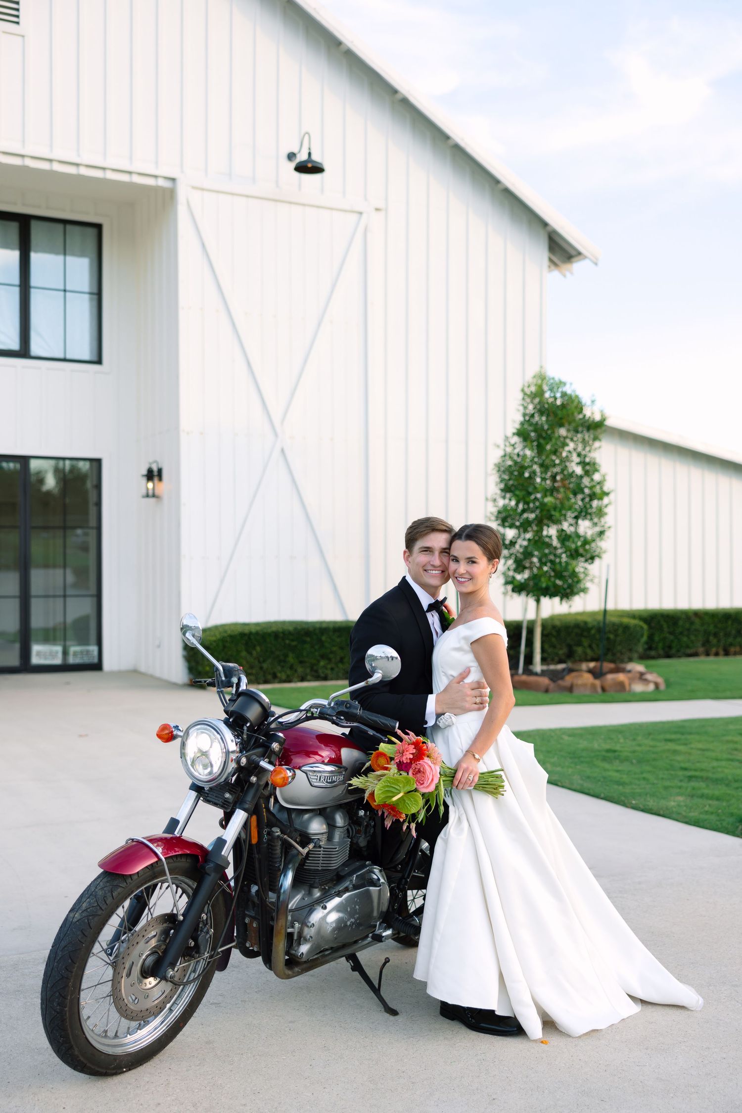 Bride and groom posing with a vintage red motorcycle in front of a white barn wedding venue.