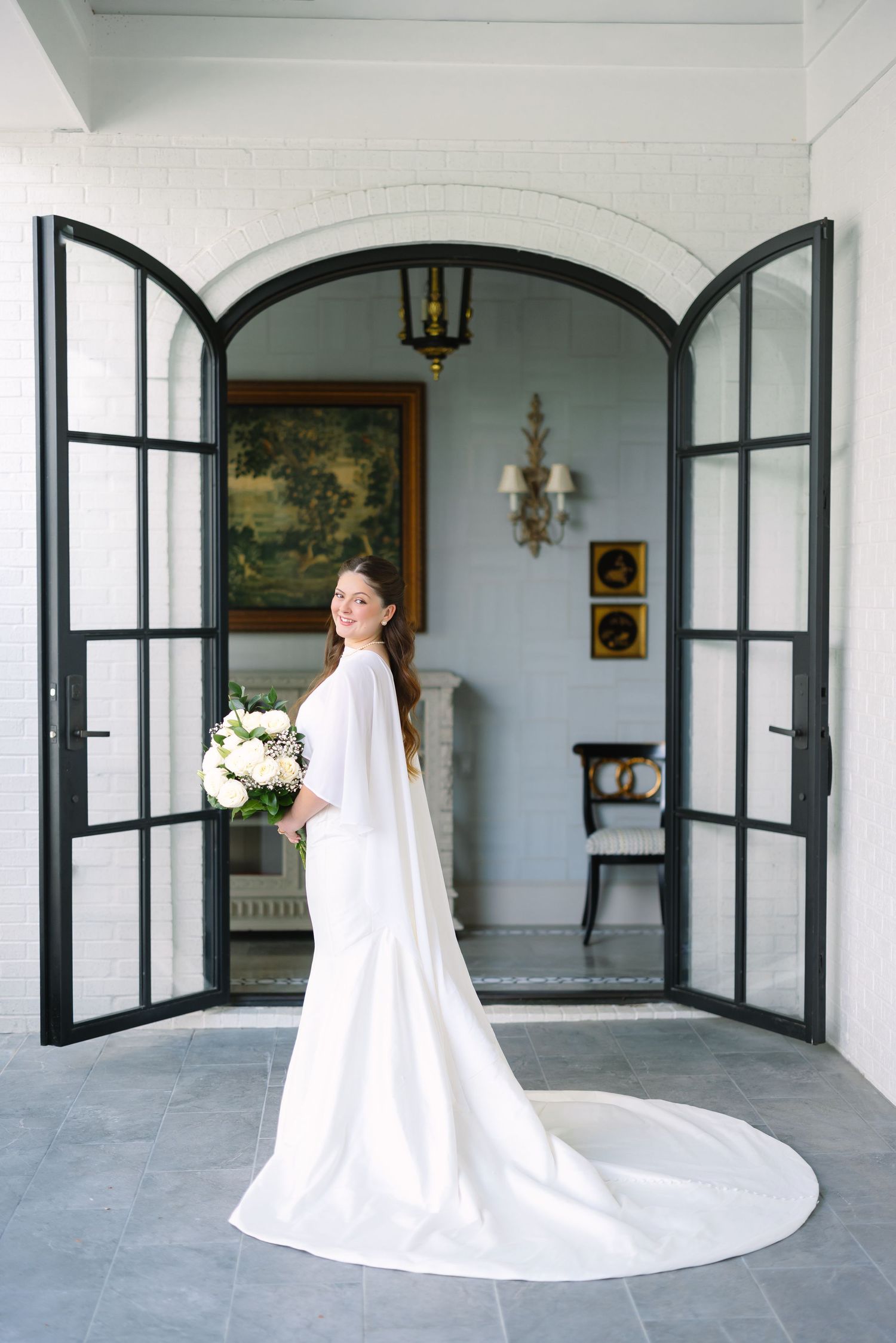 Bride in elegant white gown with train holds bouquet, standing in arched French doors of a classic venue.