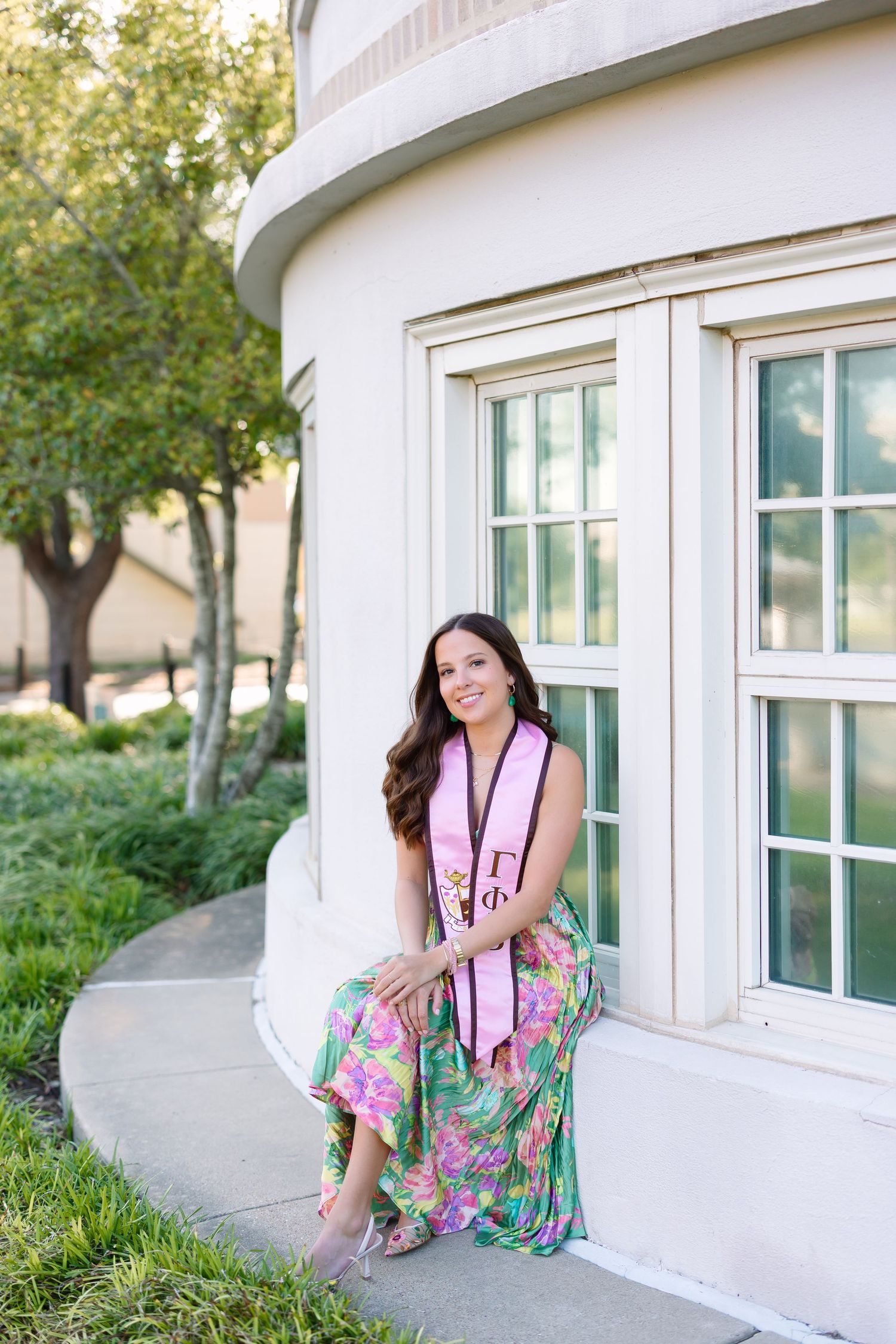 Young woman in floral dress wearing Greek sorority graduation stole sits outside university building smiling.