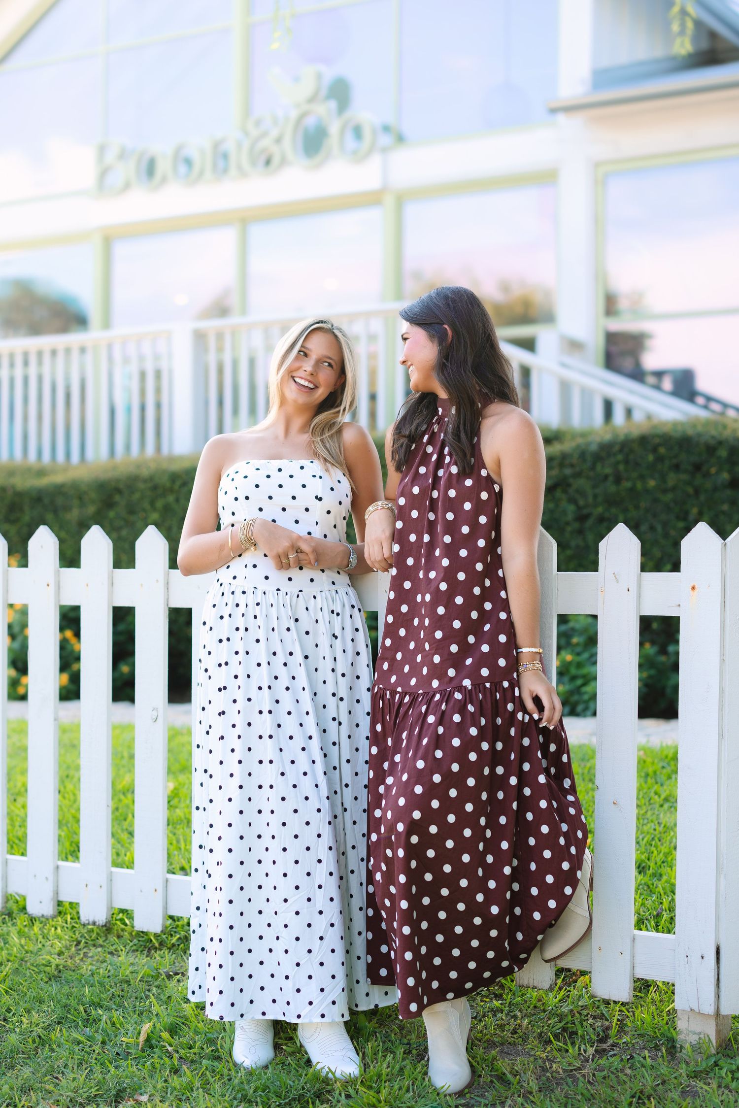 Two people in polka dot dresses stand by a white picket fence.