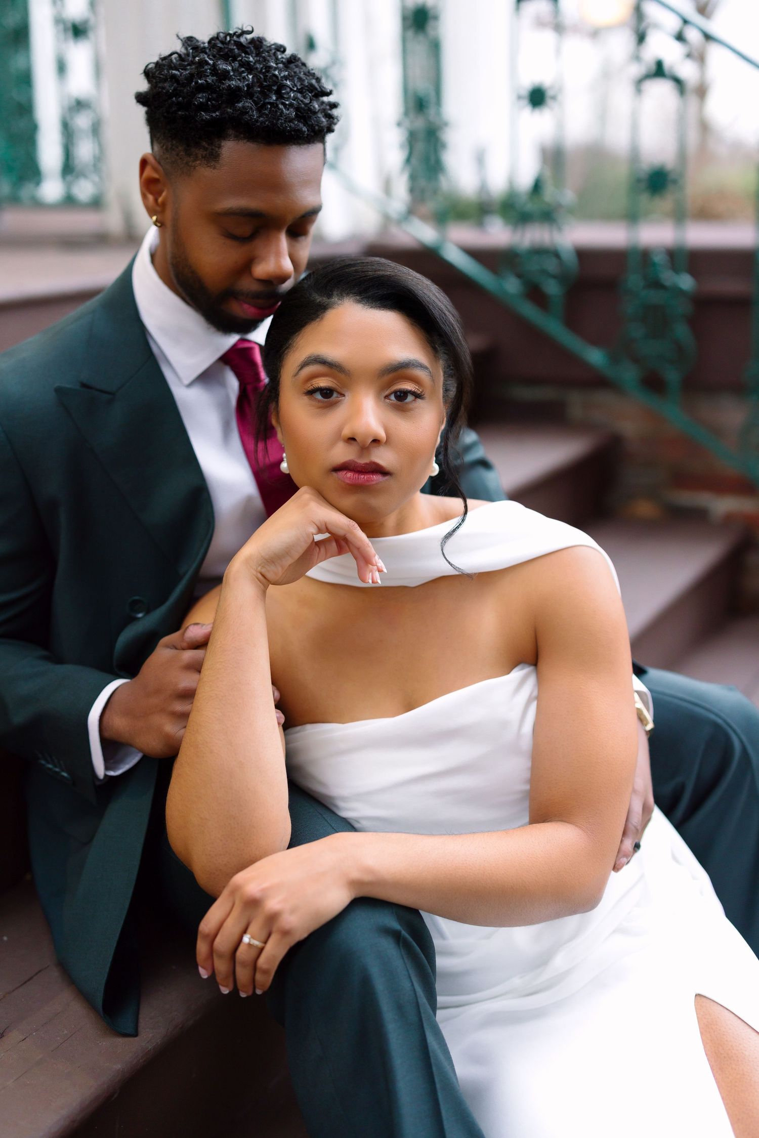 Bride in white gown sits with groom in dark suit embracing her on outdoor steps at wedding venue.
