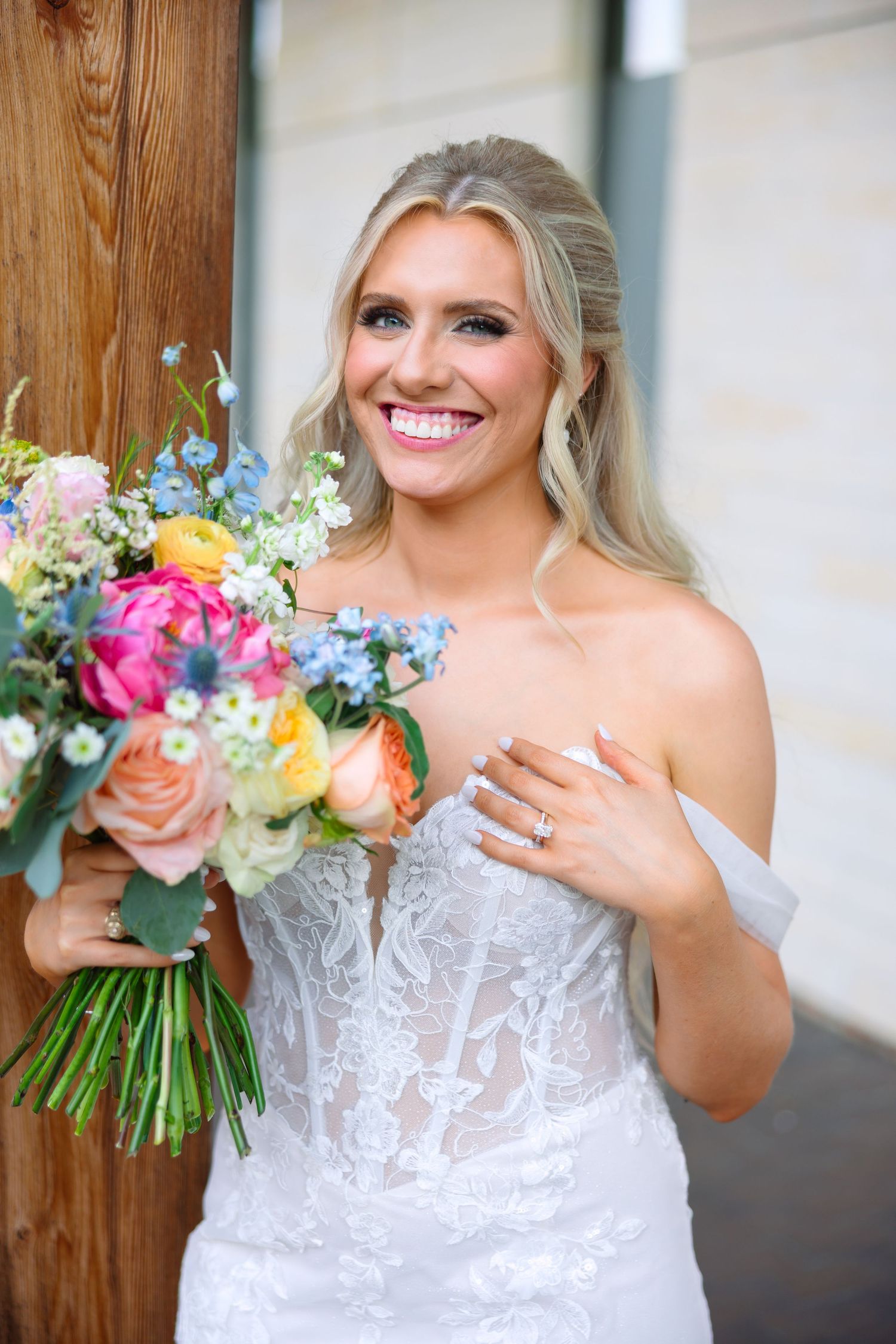 Smiling bride in lace off-shoulder gown holds colorful spring bouquet with pink, orange, and blue flowers.