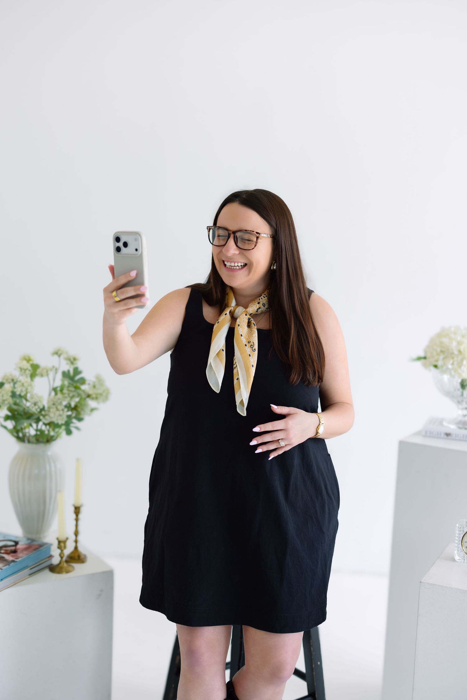Pregnant woman in black dress taking selfie, wearing glasses and yellow scarf, smiling in bright white room.
