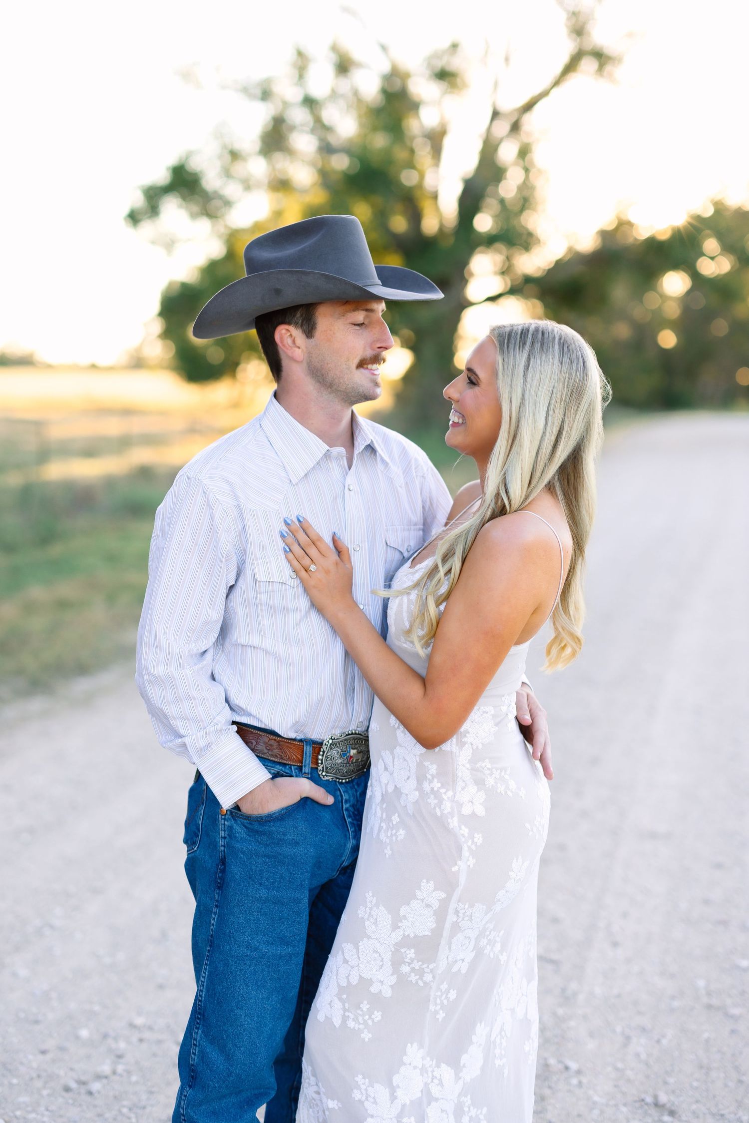 Couple in golden hour light, man in cowboy hat and jeans, woman in floral lace dress, standing on a dirt road.
