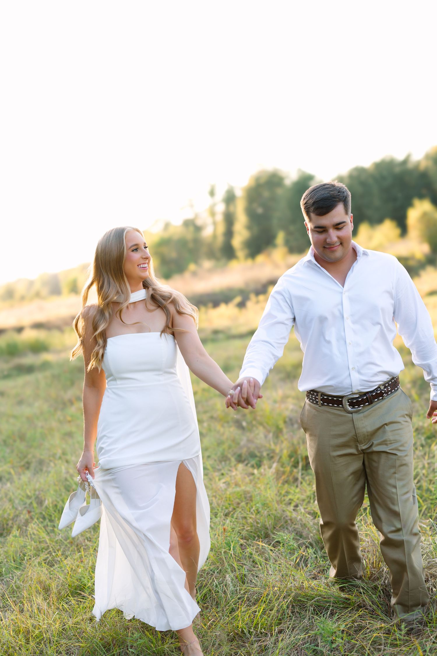 Couple holding hands walking through a sunny green field, woman in white dress carrying shoes, man in white shirt and khakis.