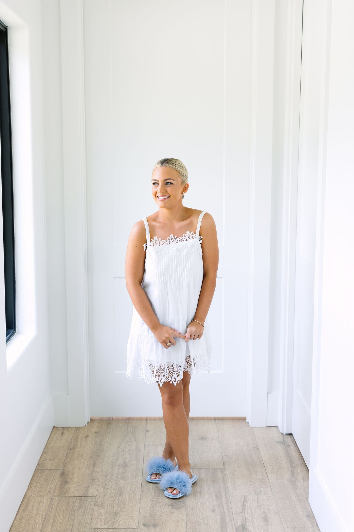 Bride-to-be wearing white lace mini dress and blue fluffy slippers stands smiling in bright white doorway.