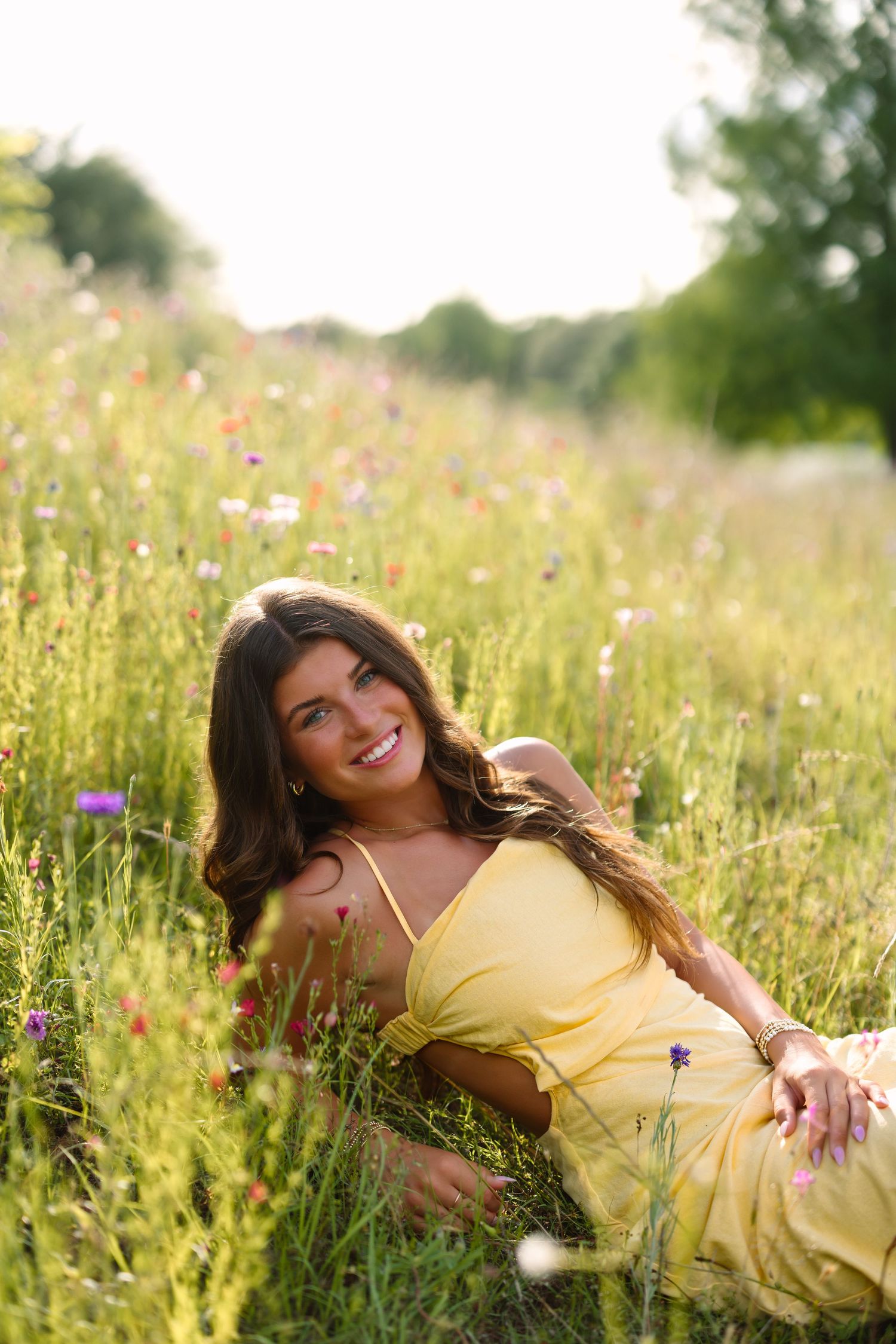 A smiling woman in a yellow dress sitting in a sunny wildflower meadow with colorful blooms and green grass.