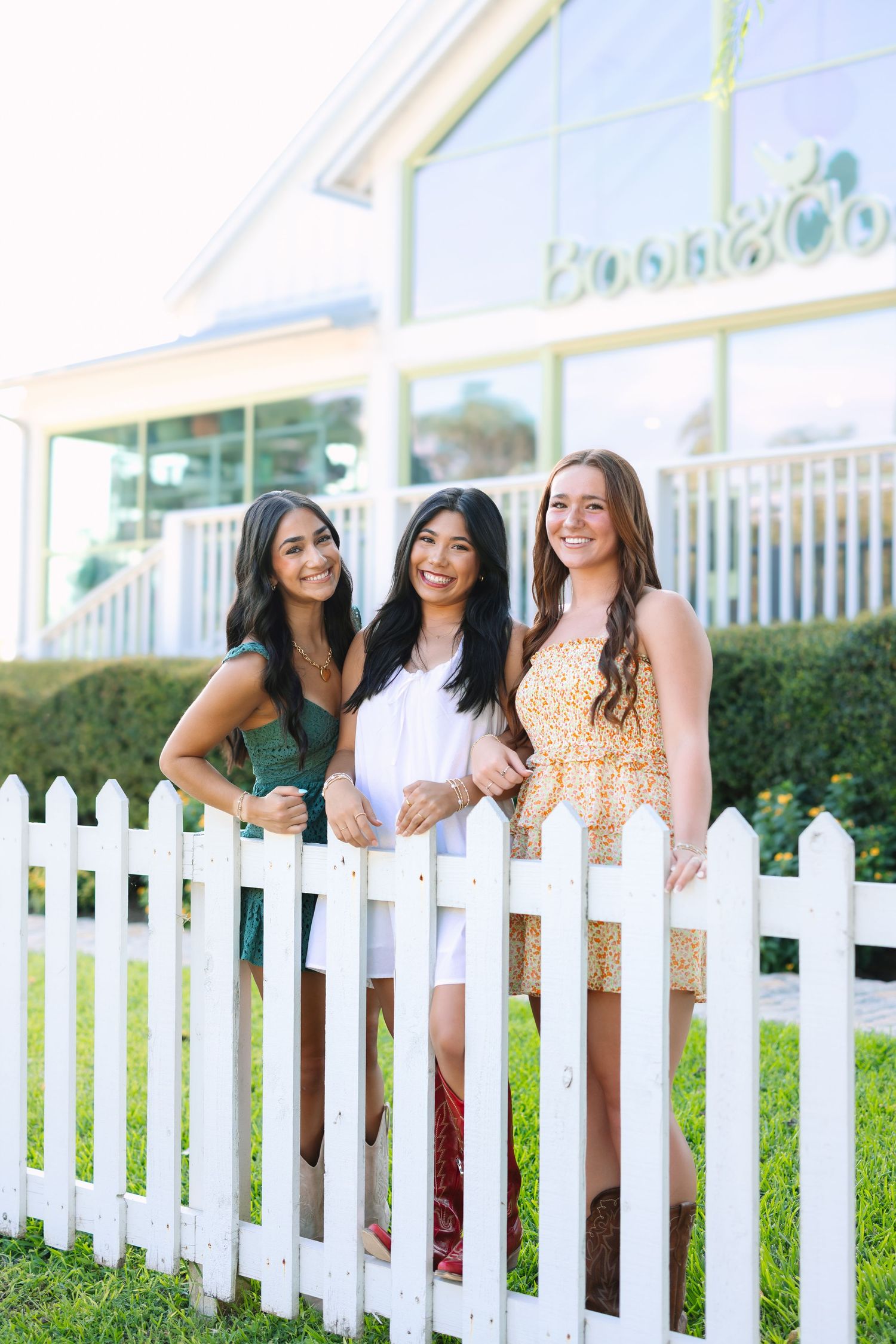Friends pose playfully by a white picket fence on a sunny day.
