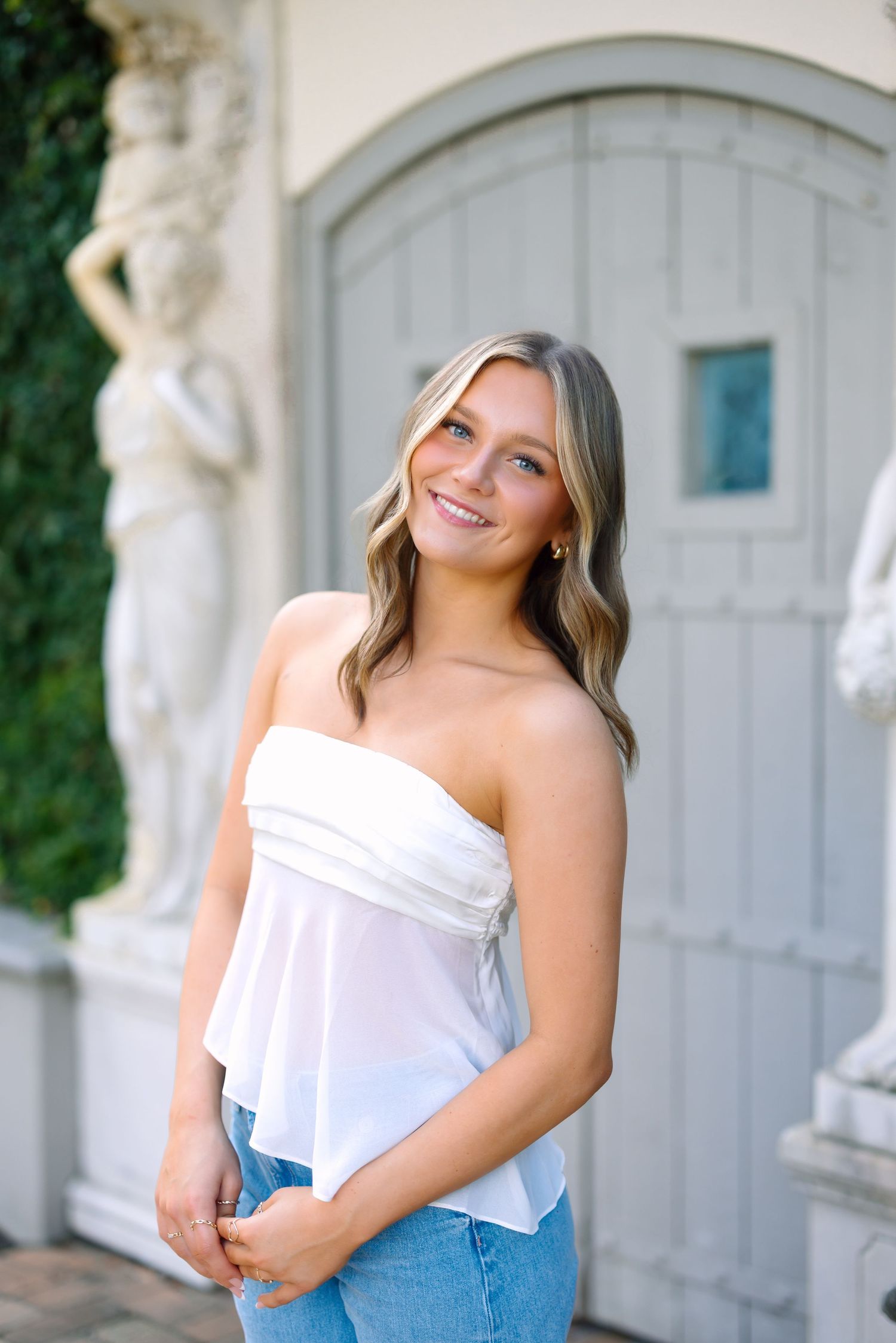 Young woman smiling in white strapless top and jeans, posing outdoors near elegant stone statues and gray door.
