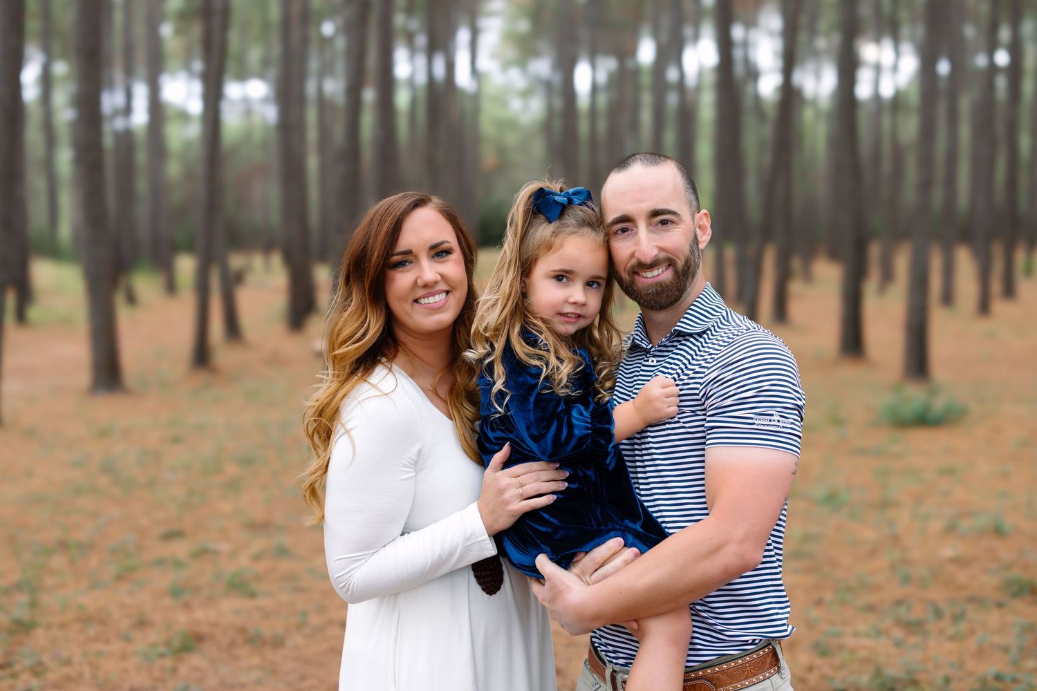 Family of three smiling in a pine forest, mother in white dress, father in striped shirt, holding young girl in blue dress.