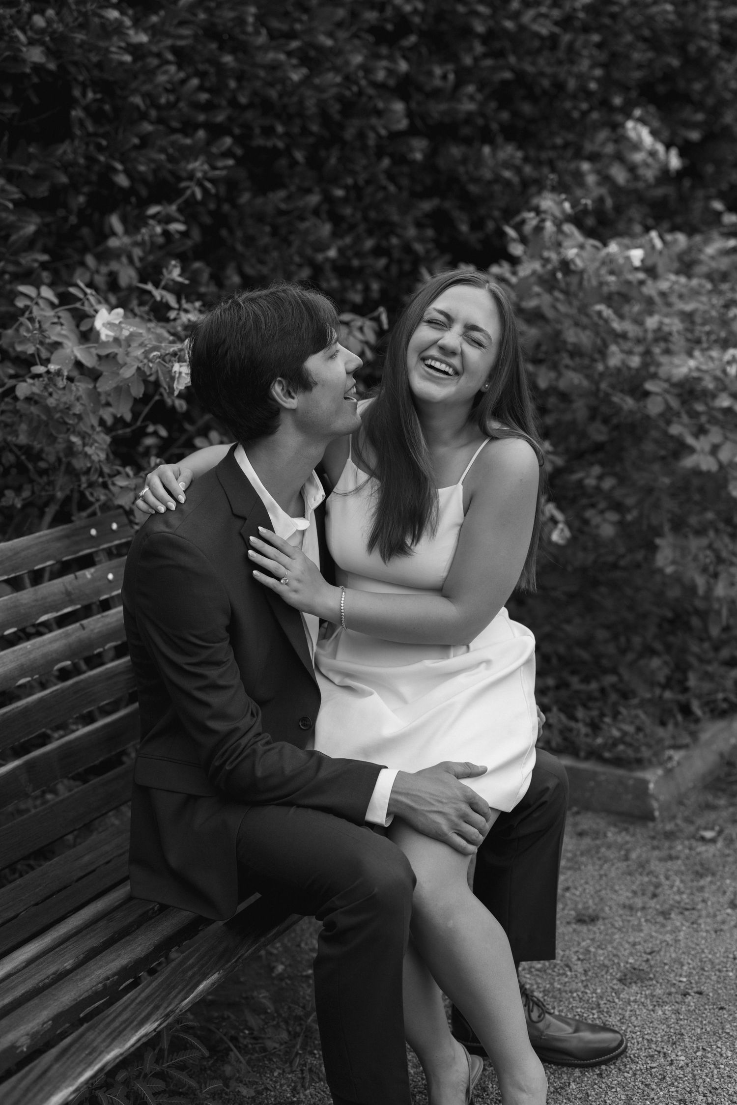 A couple shares joyful laughter while sitting together on a wooden bench in a garden setting.