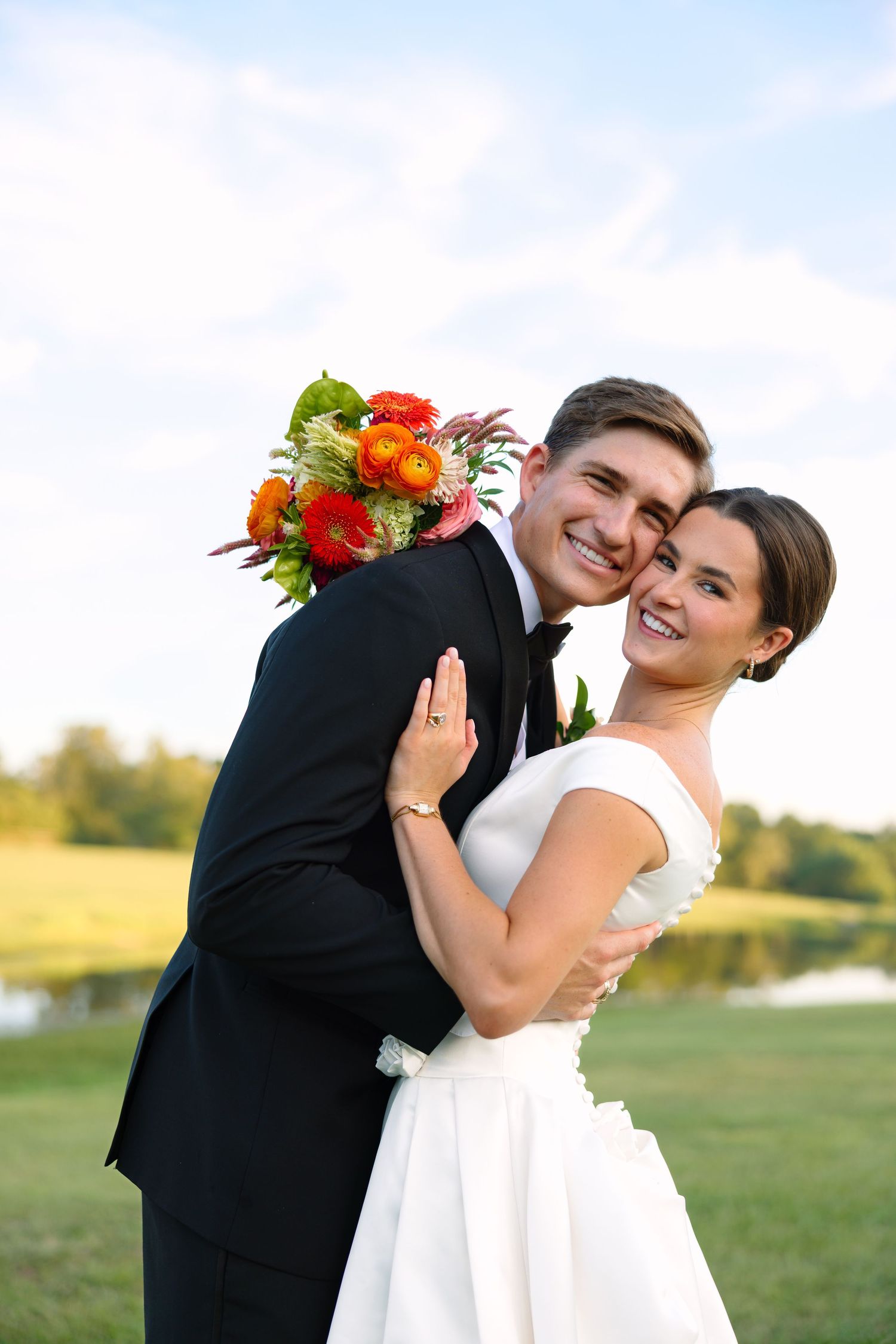 Happy bride and groom embrace outdoors holding colorful orange and red bouquet with scenic green landscape.