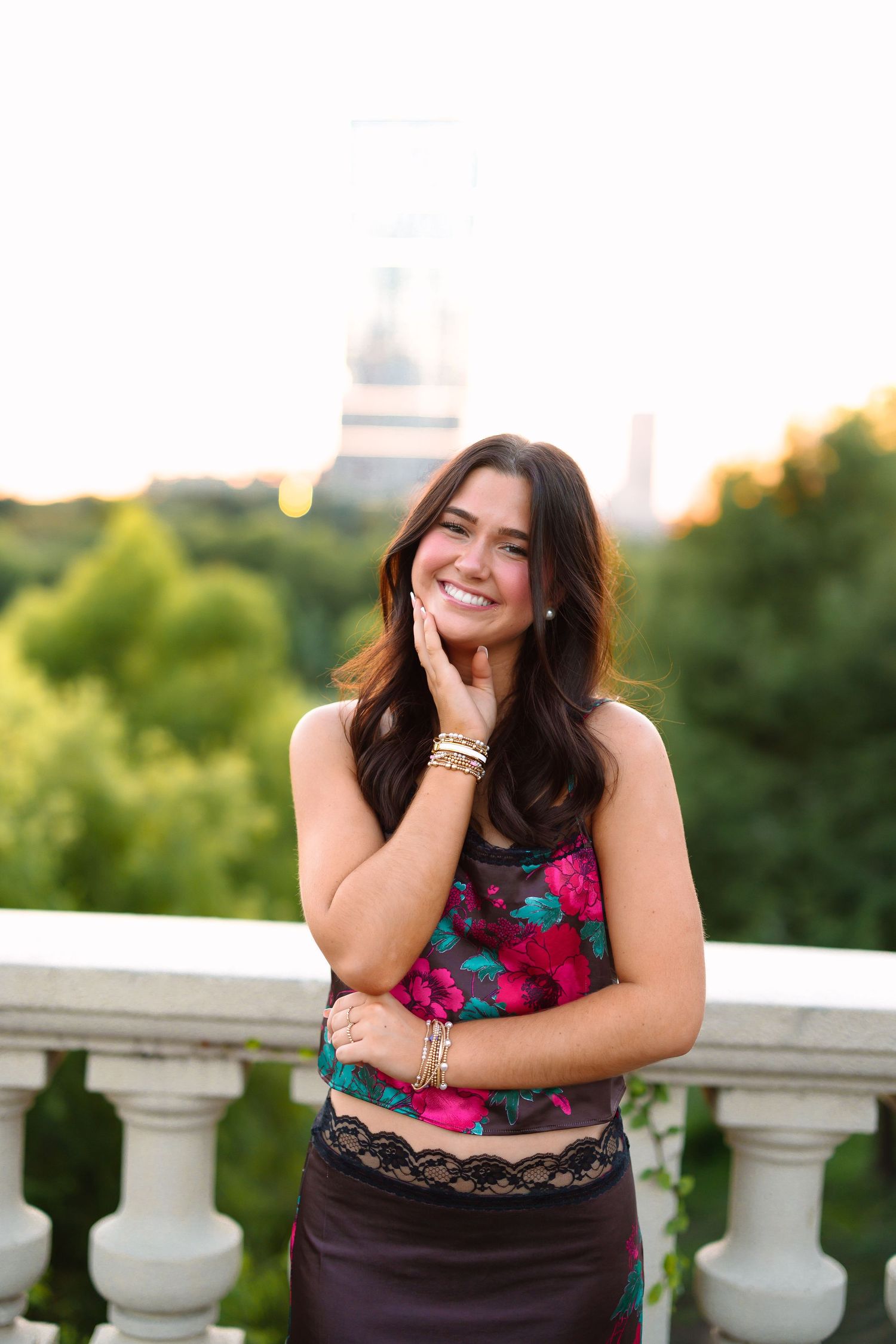 Woman in a floral dress poses joyfully against an outdoor balcony railing at sunset.