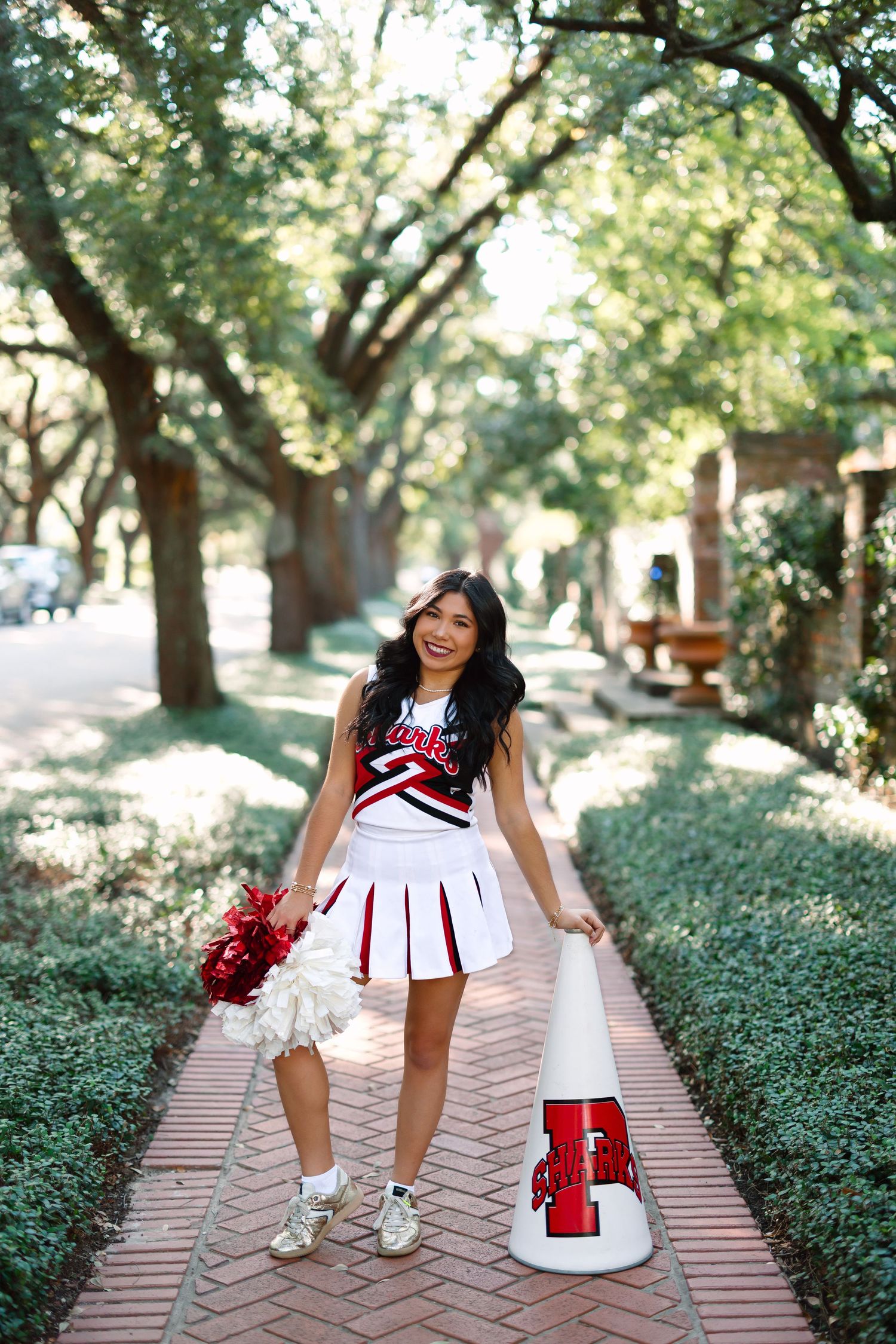 Cheerleader poses with pom poms and megaphone on brick pathway lined with trees.