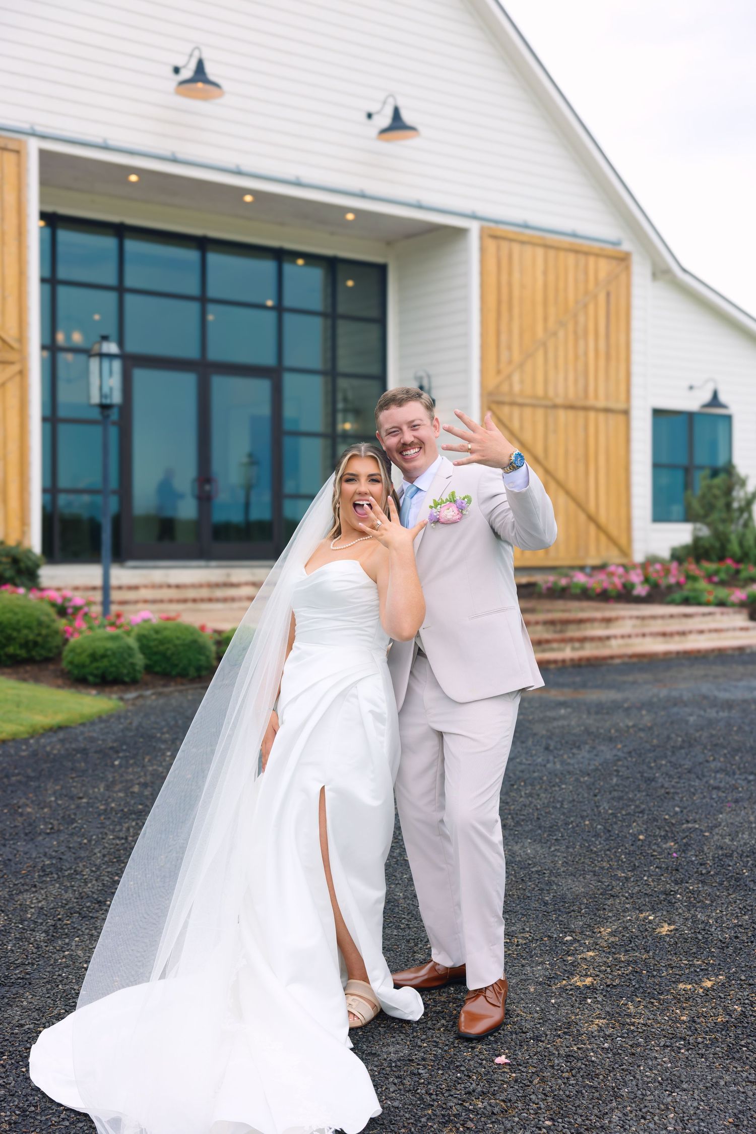 Joyful bride and groom waving in front of a white barn wedding venue, bride in white gown with veil.