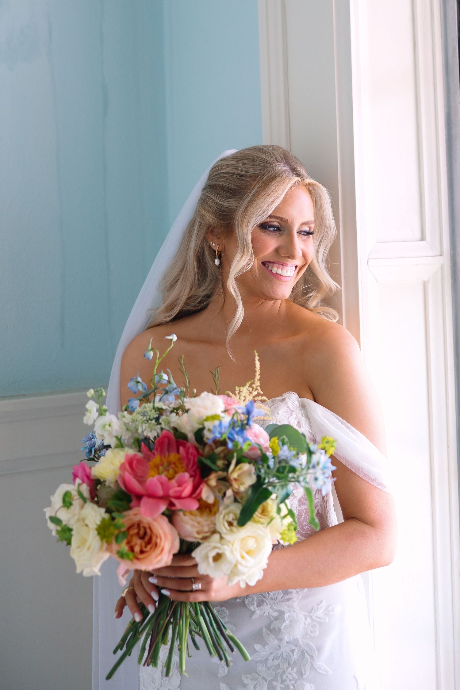 Smiling bride in strapless lace gown holds vibrant bouquet of pink, white, and blue flowers by window.
