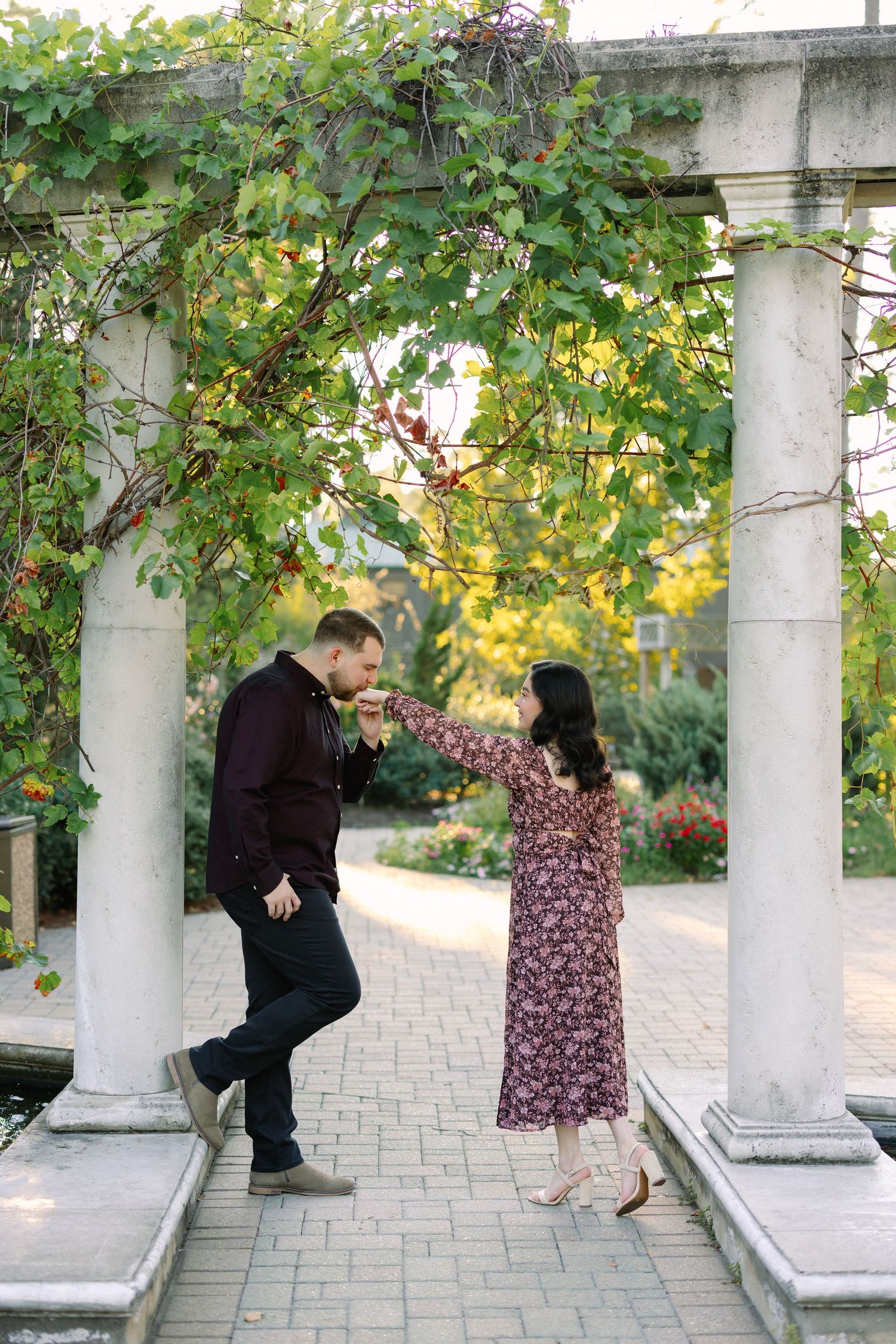 A couple shares a romantic moment under a vine-covered pergola in a lush garden, man kissing woman's hand.