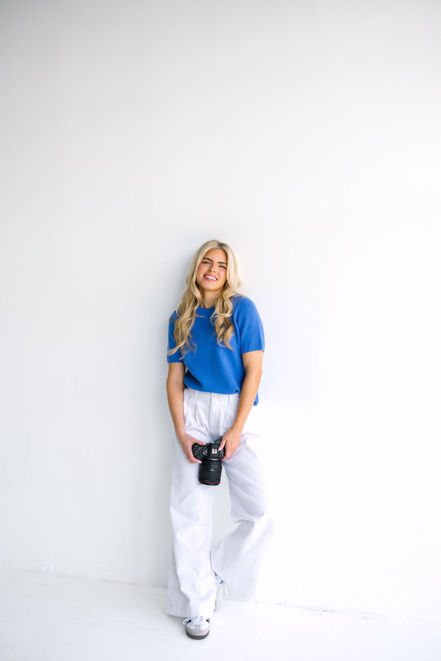 Woman in bright blue t-shirt and white pants standing against white wall holding camera.