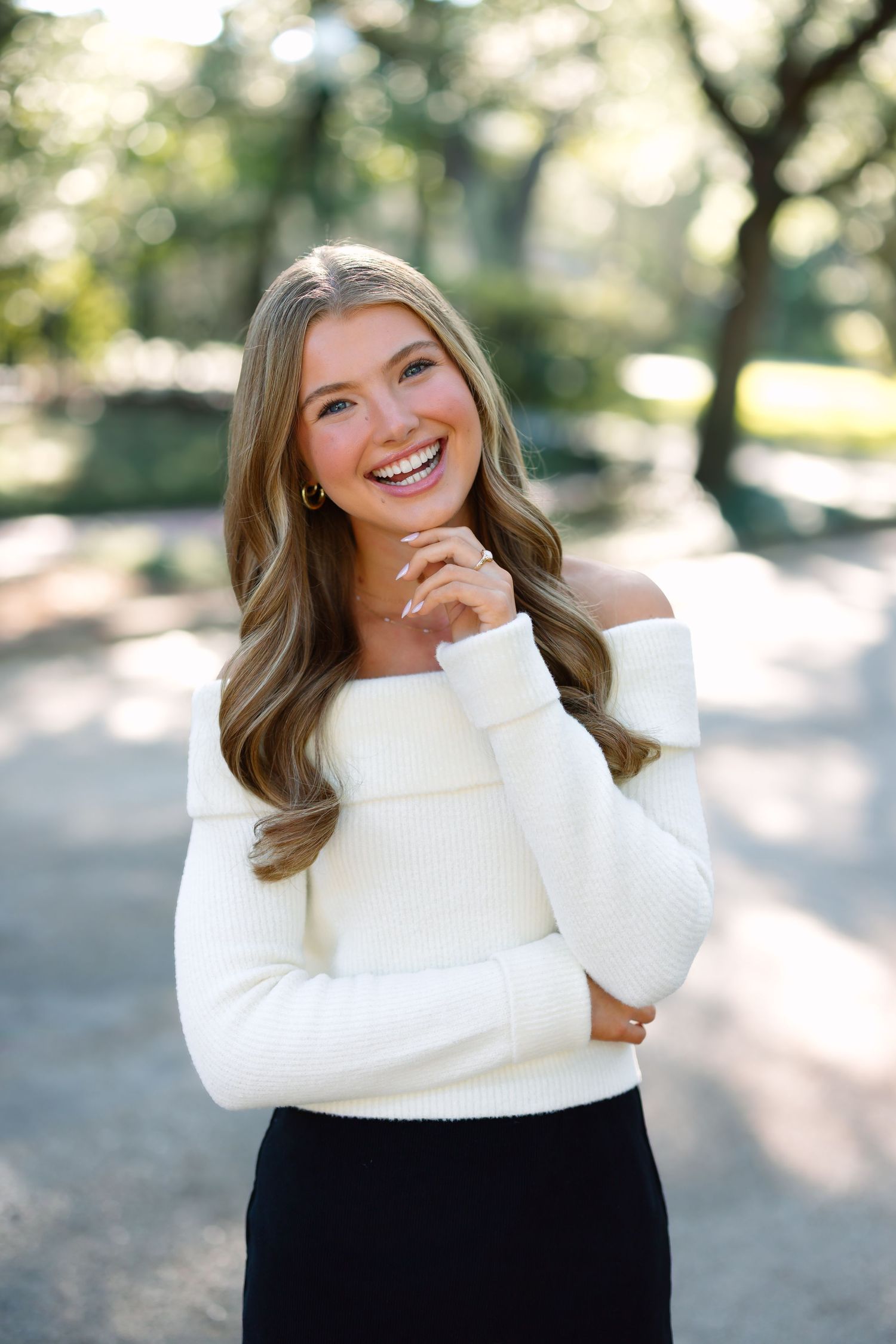 Young woman smiling outdoors in white off-shoulder sweater and black skirt, with long wavy hair, in a park setting.