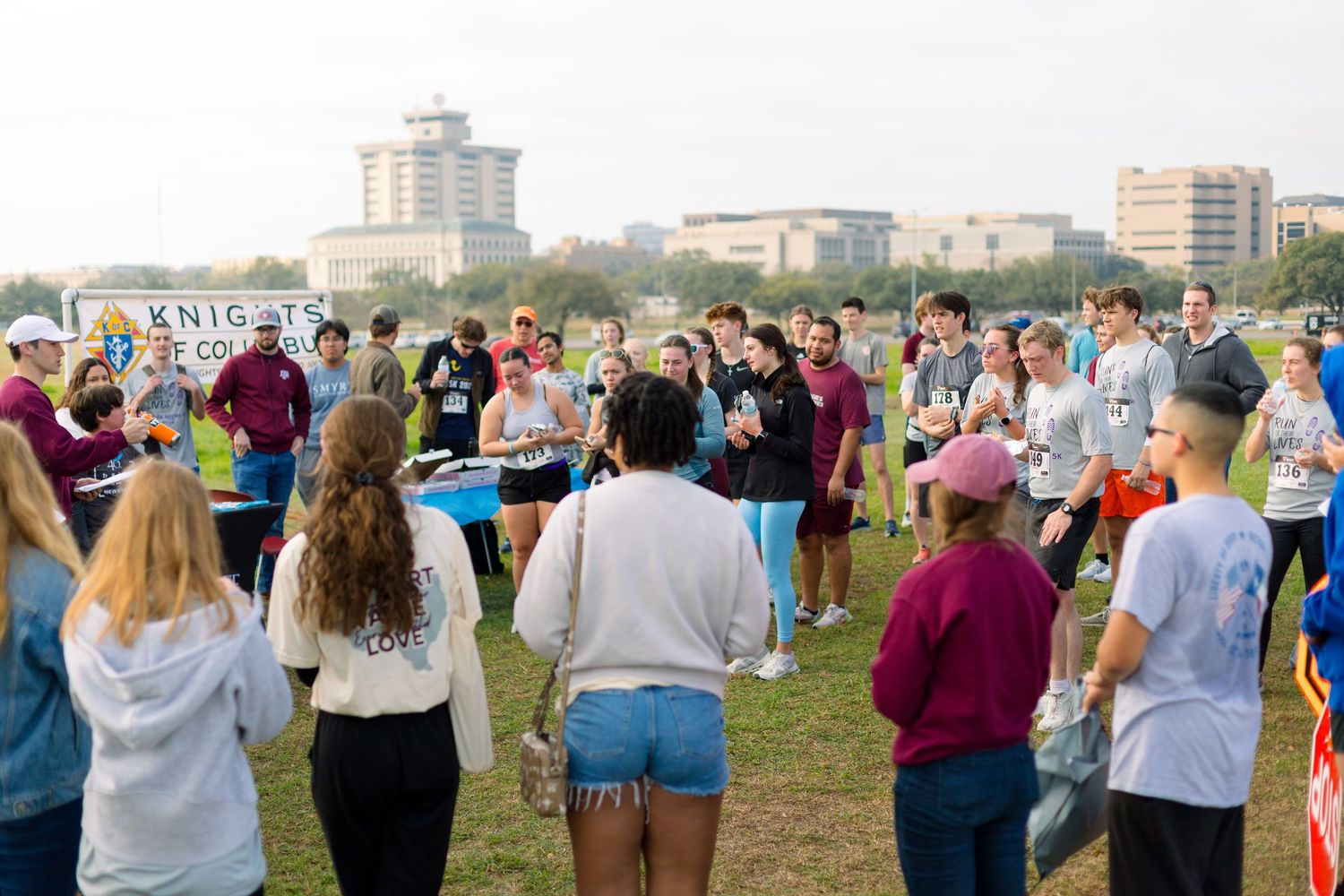 Crowd of people gathered on a grassy field with buildings visible in the background.