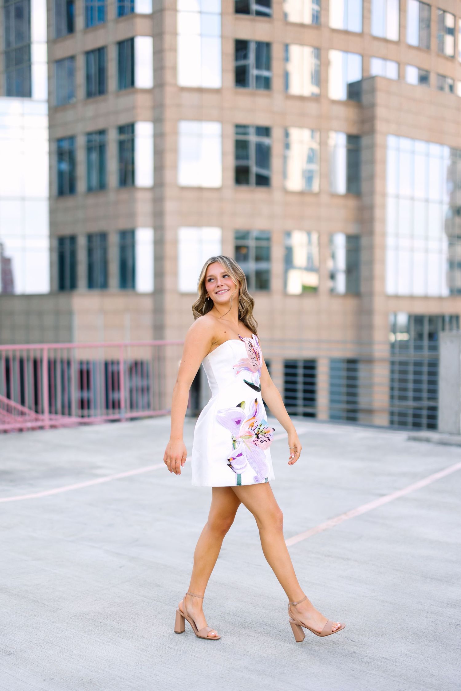 Woman in white floral strapless mini dress and nude heels walking on rooftop with city buildings in background.