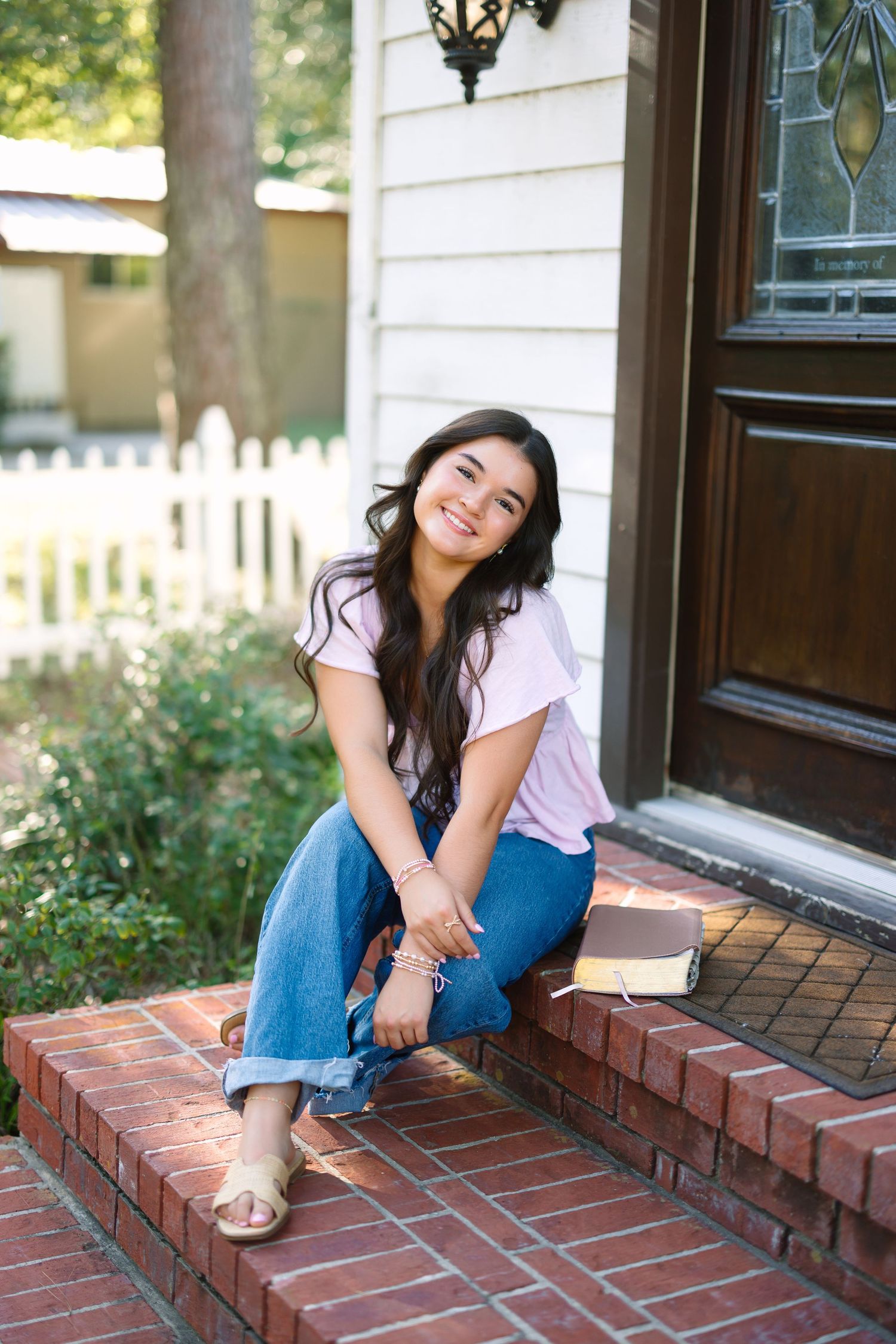 Smiling young woman in pink top and jeans sitting on brick porch steps with a book beside her.