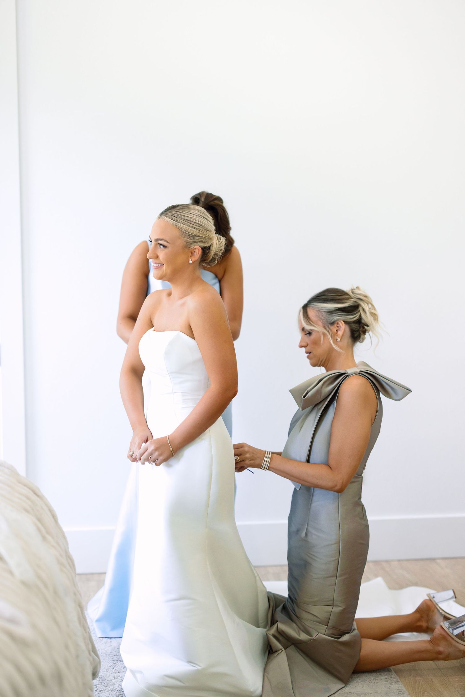 Bride in strapless white wedding gown gets dress fastened by bridesmaid in gray dress during bridal preparations.
