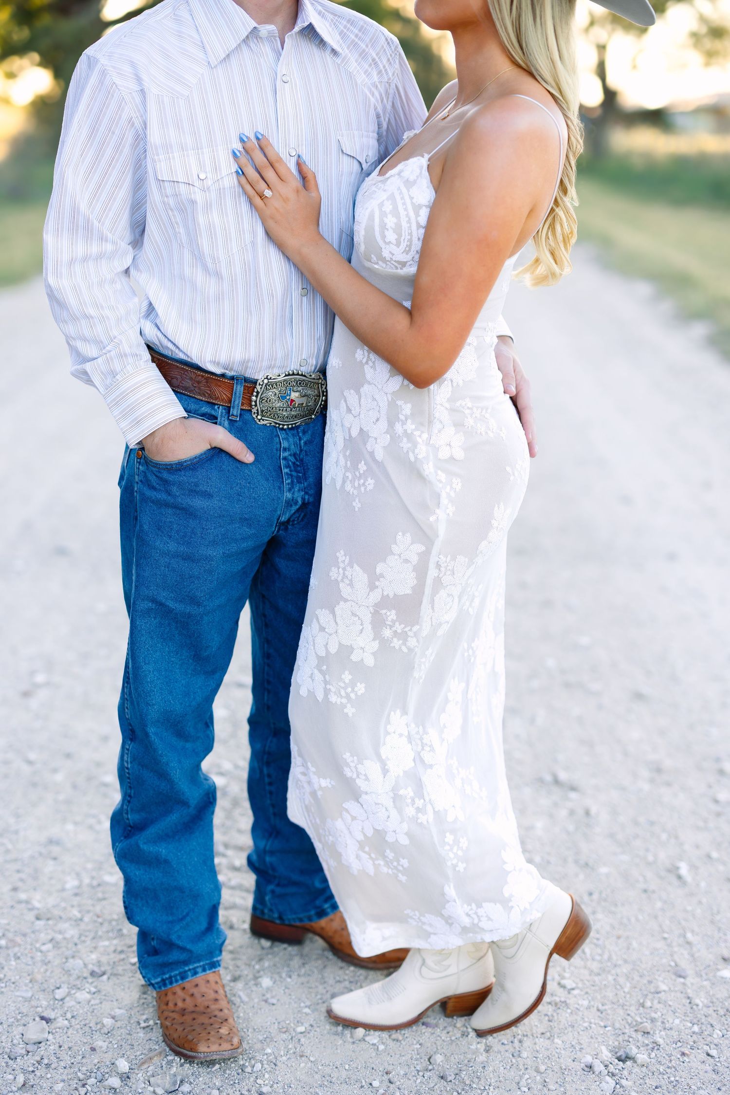 Close-up of couple in Western attire, man with decorative belt buckle and cowboy boots, woman in white lace dress.