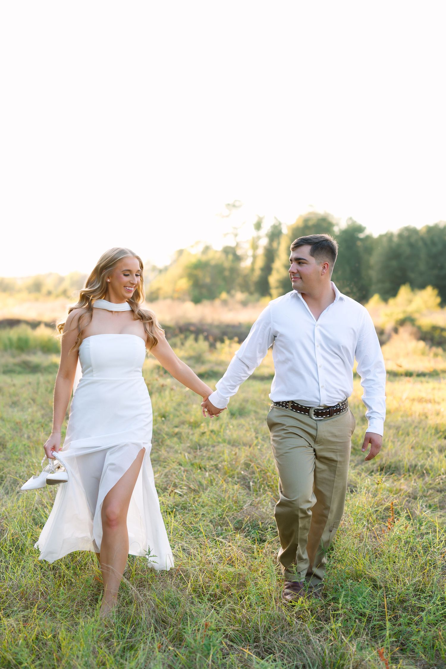 Couple holding hands walking through a sunny field, woman in white dress carrying shoes, man in white shirt and khakis.