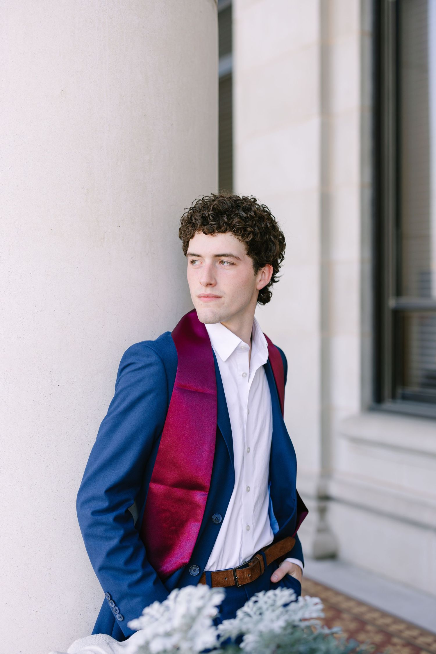 A person in a navy blue blazer and burgundy scarf stands against a white column background.