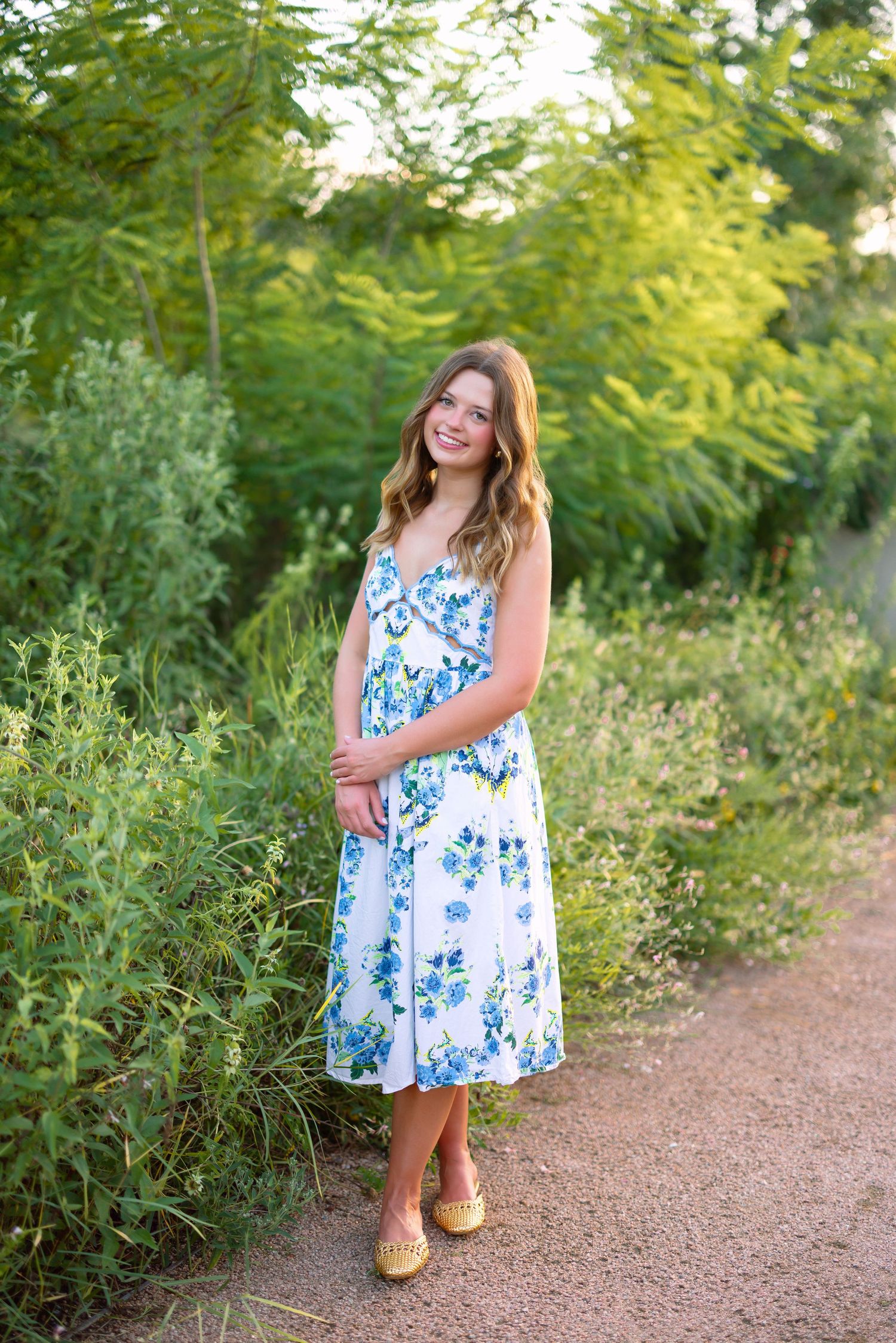 Young woman in white floral dress smiling on a dirt path surrounded by lush green trees and plants.