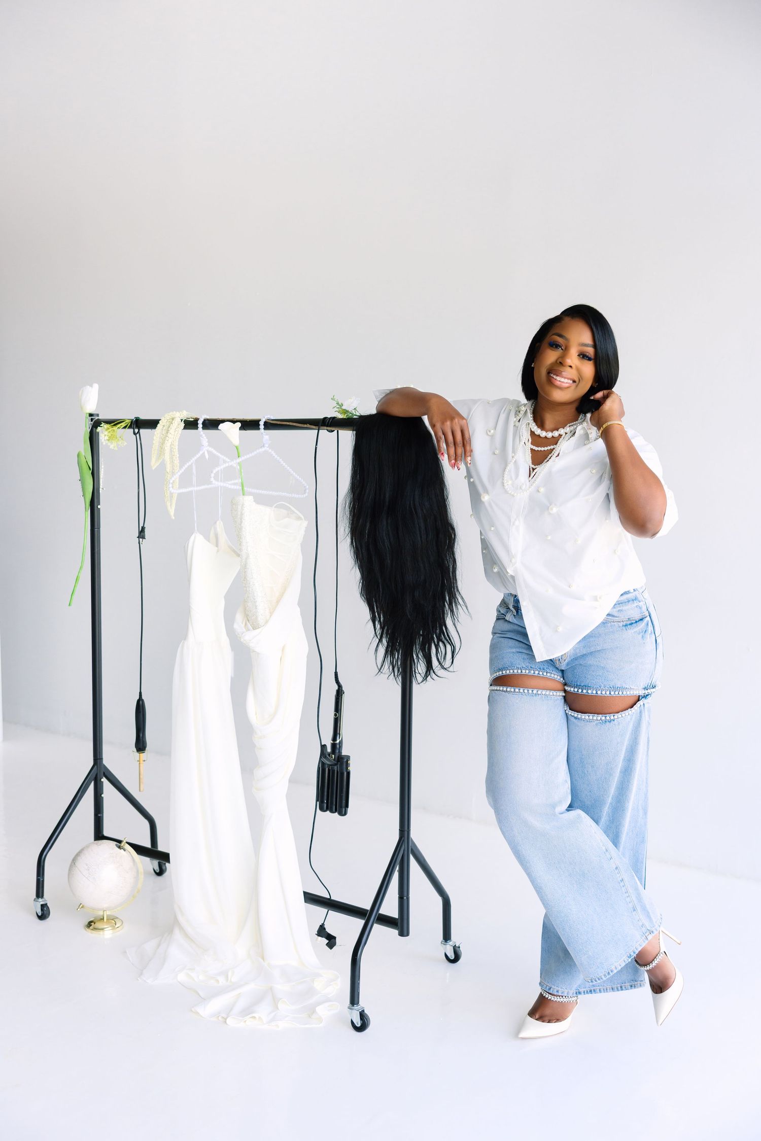 Smiling woman in white blouse and ripped jeans posing by a clothing rack with white dresses and a black wig.